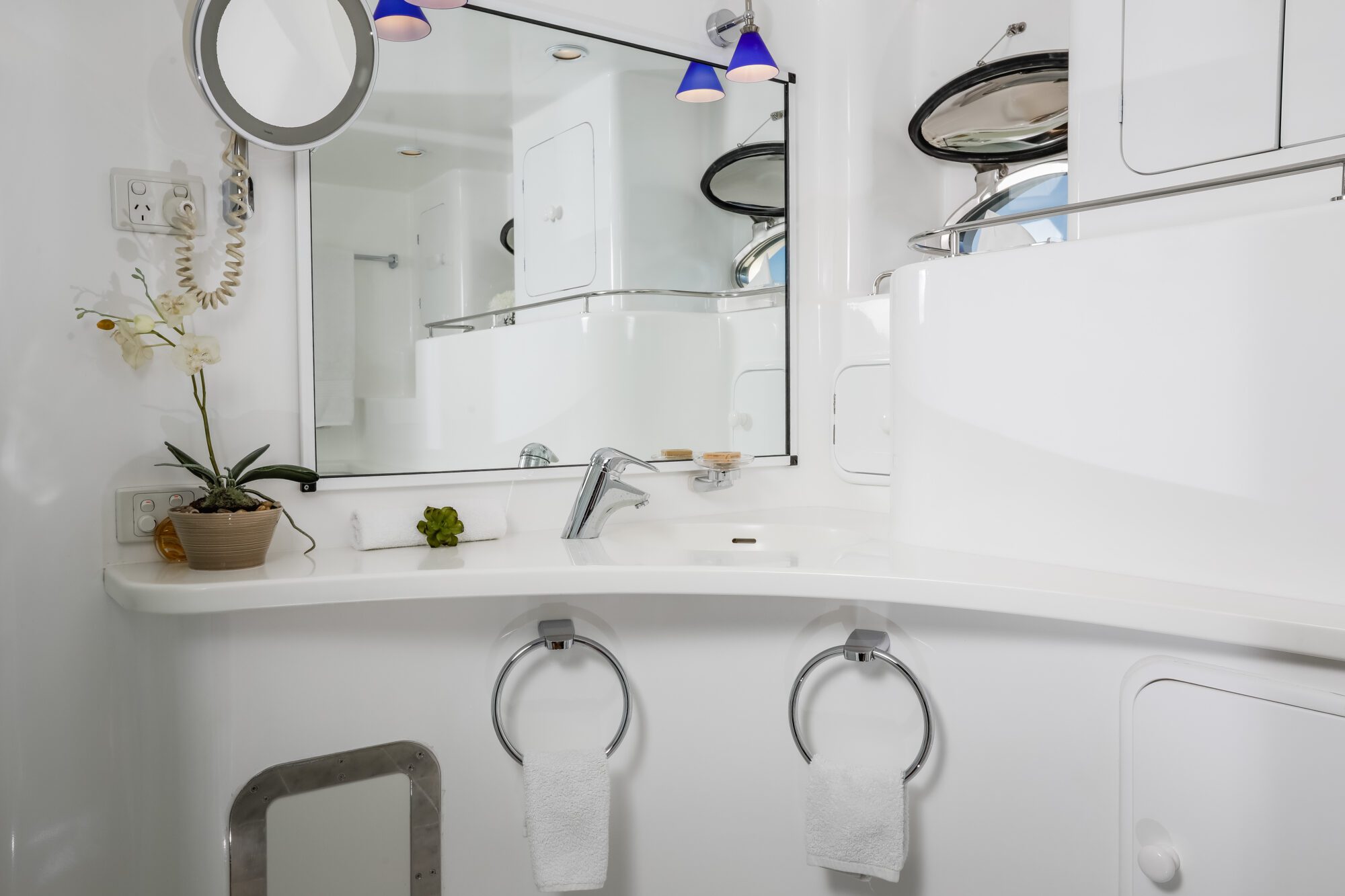 A modern white bathroom with a curved counter, two towel rings with white towels, a potted orchid, a large mirror, and blue pendant lights above the sink. A round window and built-in cabinets are also visible.