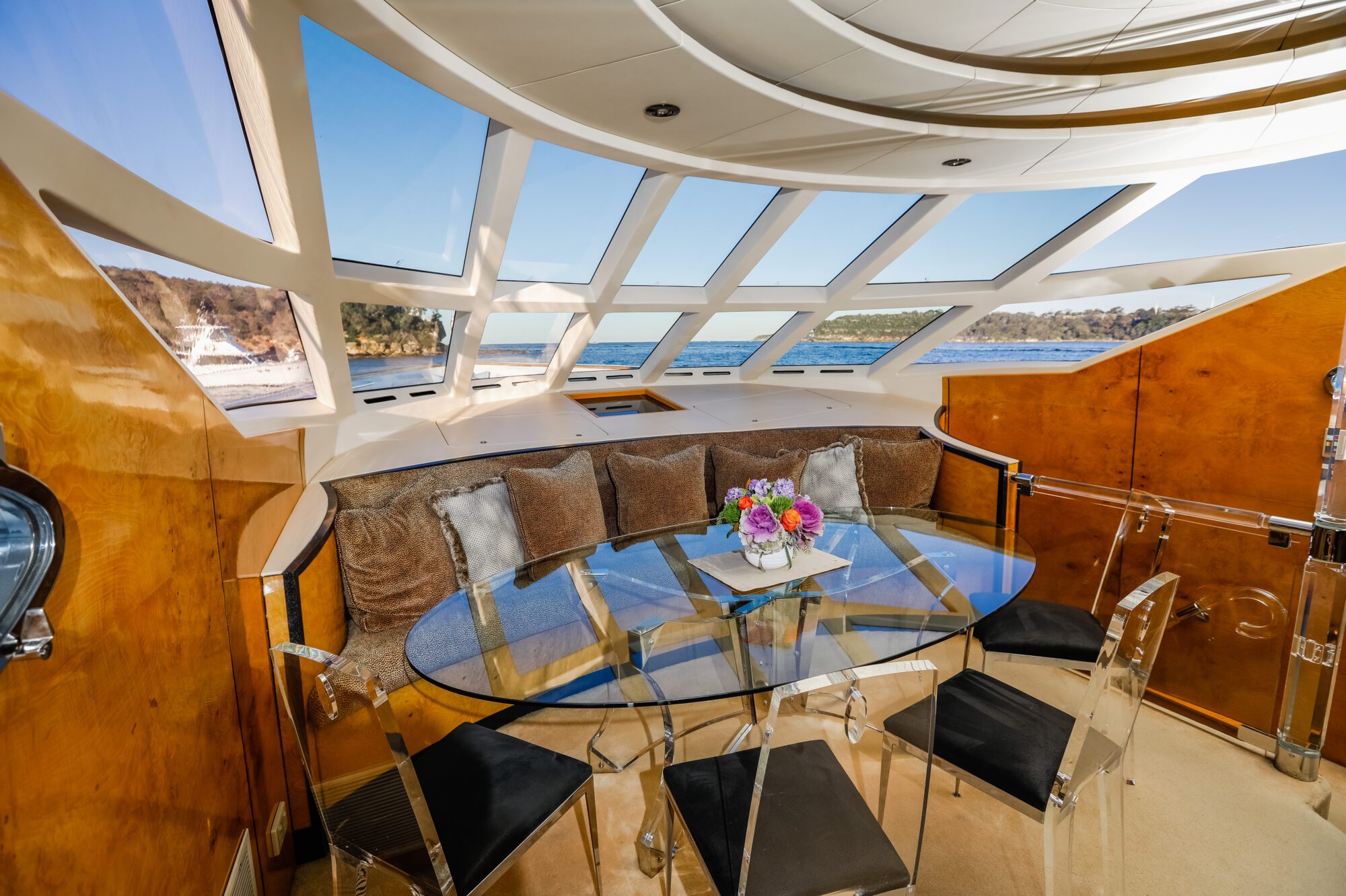 A modern yacht interior with a glass dining table, transparent chairs, a plush sofa with cushions, and large windows offering a scenic view of water and trees outside. A floral centerpiece sits on the table.