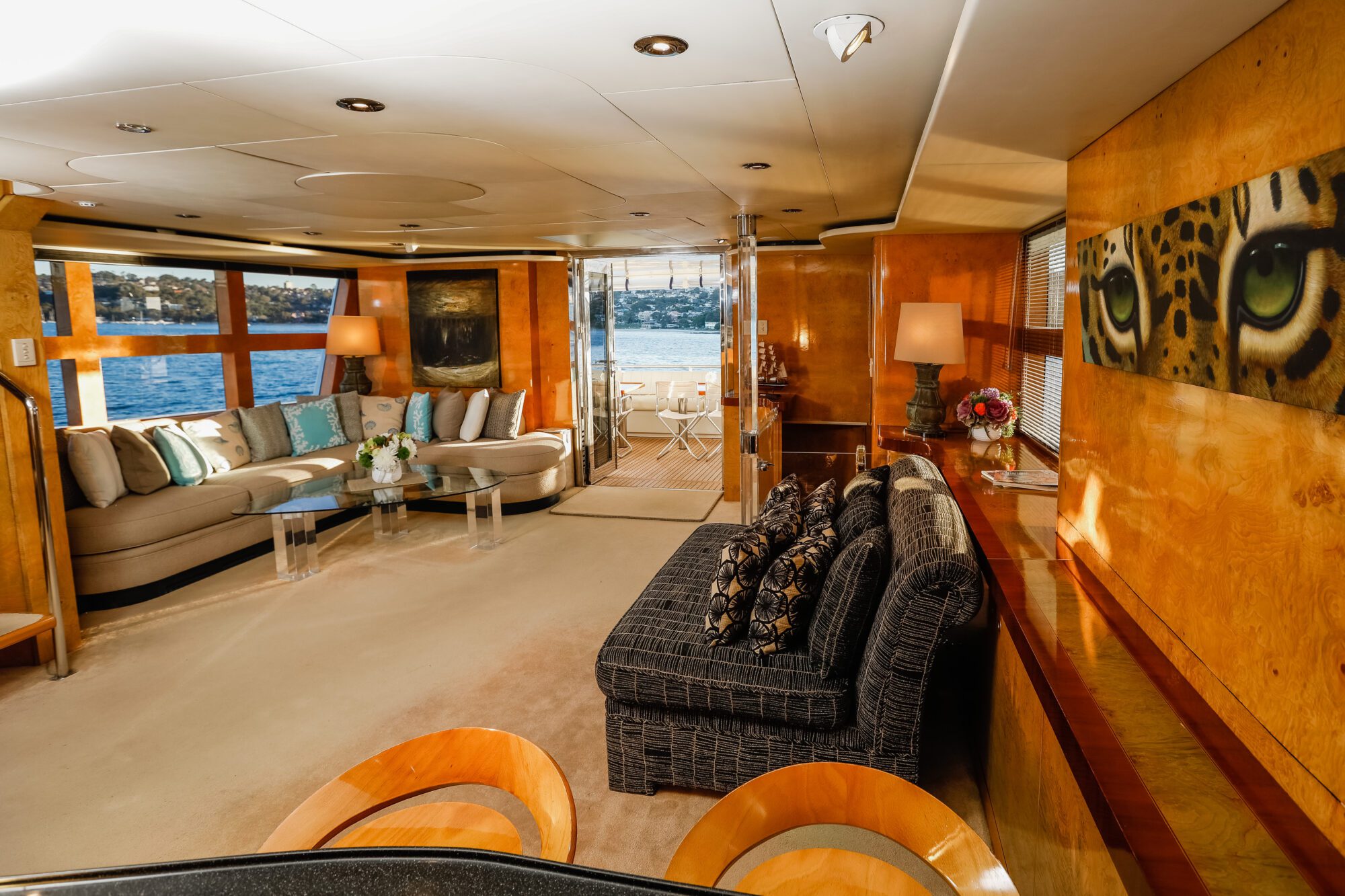 Luxurious yacht interior with plush sofas, decorative pillows, a glass coffee table, large windows overlooking the water, stylish wooden walls, and a deck area visible through glass doors at the back.