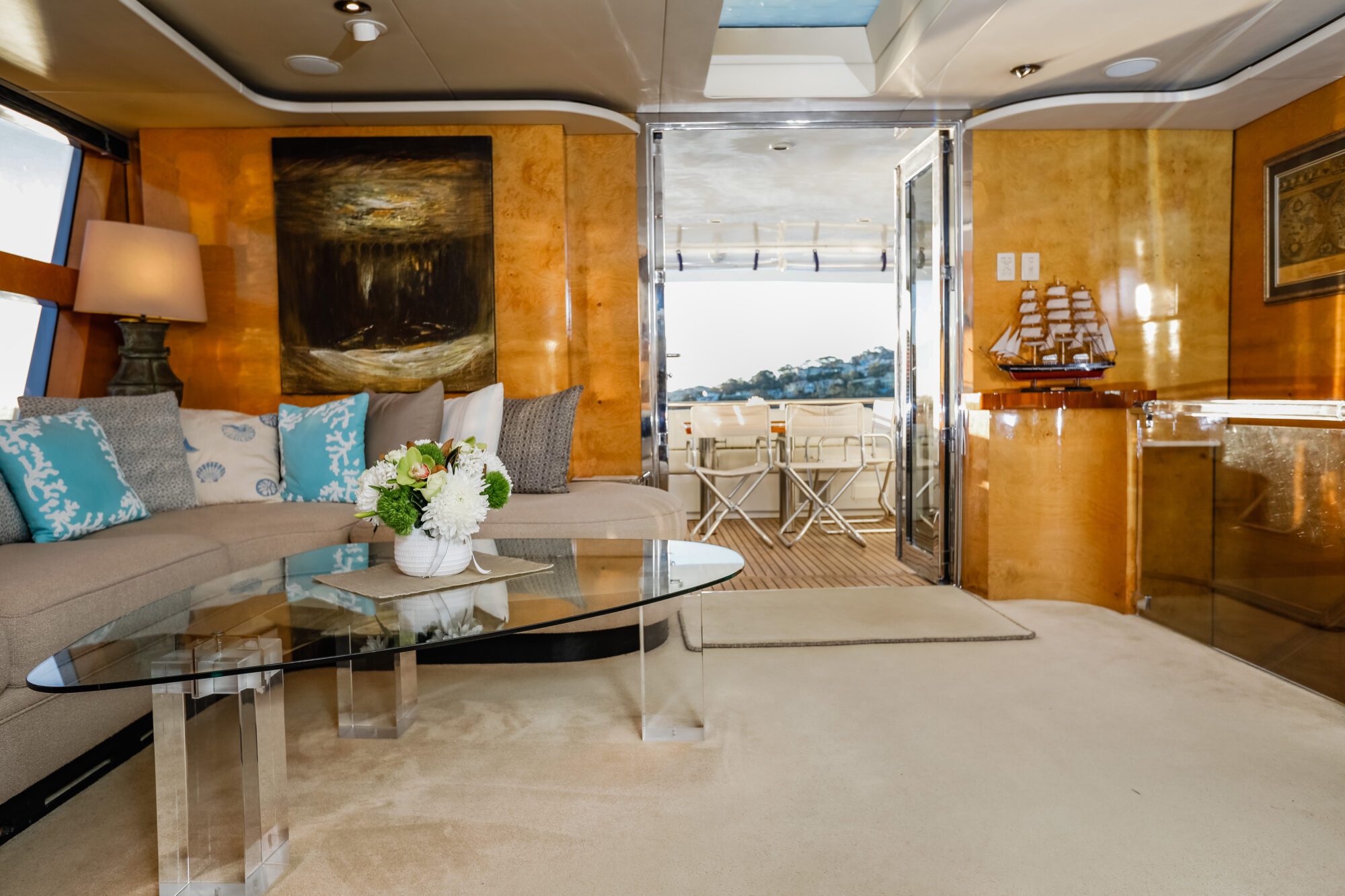 A luxurious yacht interior with a glass coffee table, beige sectional sofa with blue and white pillows, floral centerpiece, modern art on wood-paneled walls, and large windows overlooking a sunlit outdoor seating area.