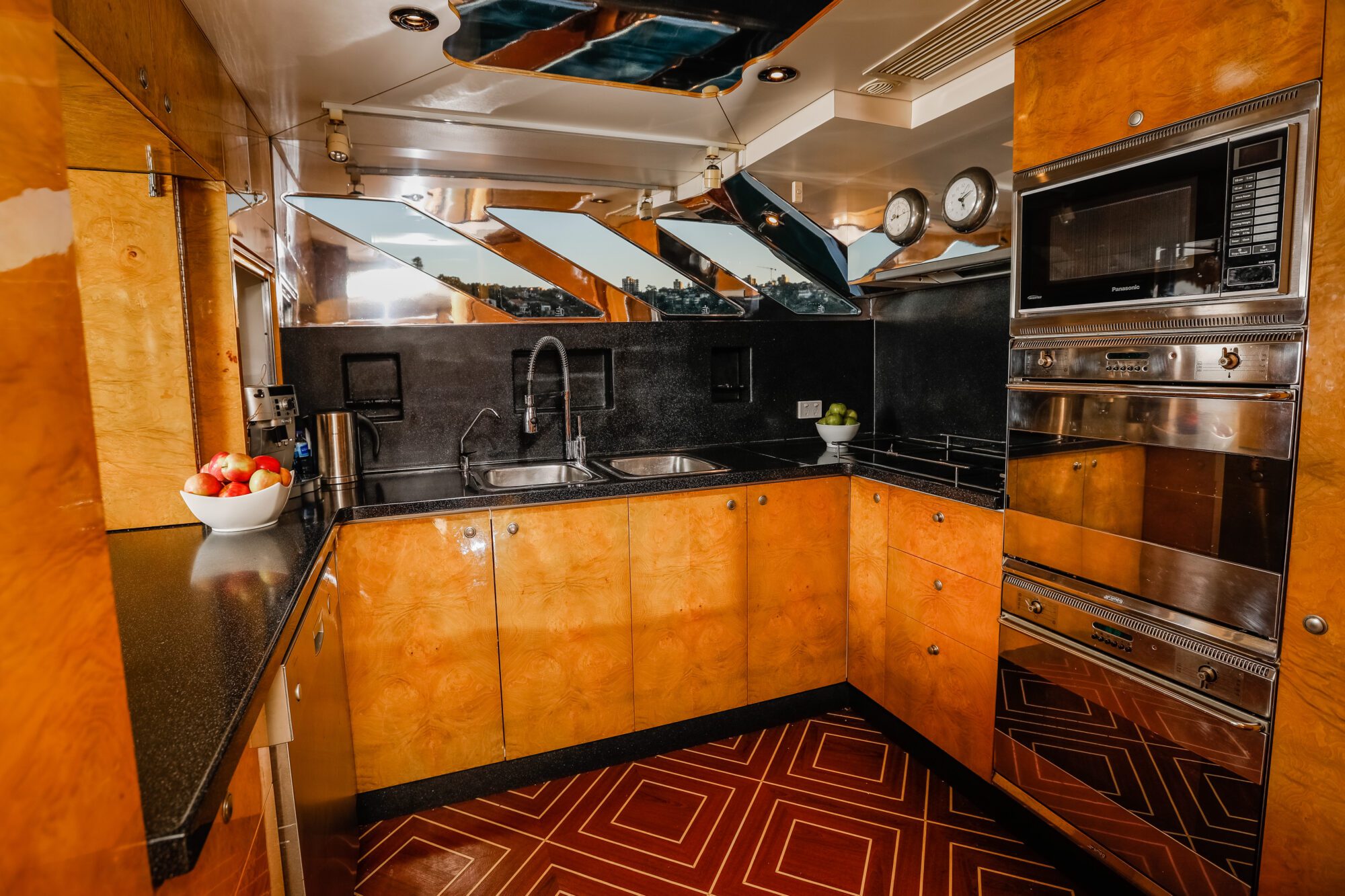 A modern kitchen with shiny wooden cabinets, black countertops, a double sink, built-in oven and microwave, mirrors on the backsplash, and a bowl of apples on the counter. The floor has a red geometric pattern.