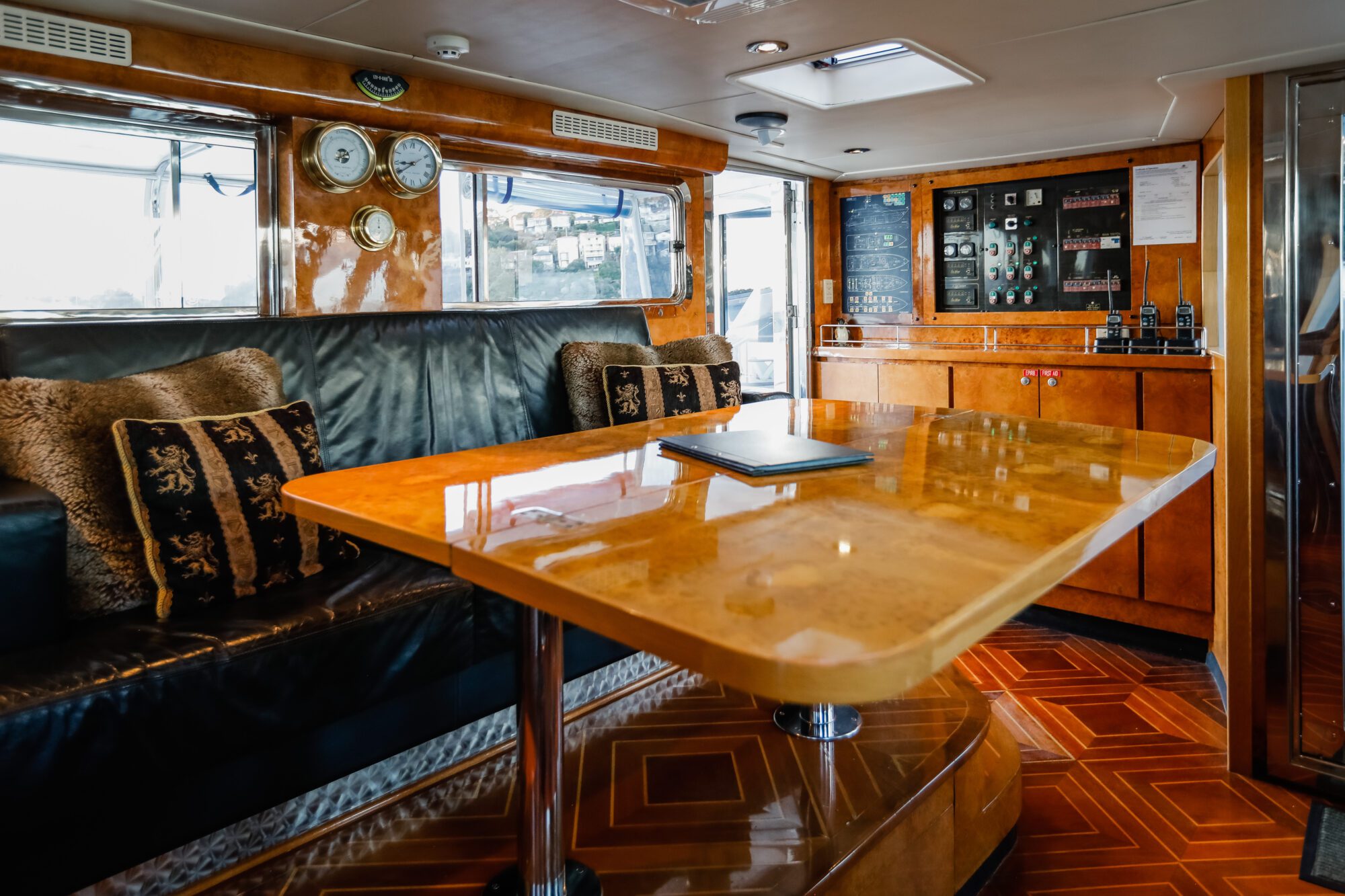 Luxurious yacht interior with a polished wooden table, black leather sofa with decorative pillows, wall clocks, and a navigation control panel, all surrounded by wood paneling and flooring.