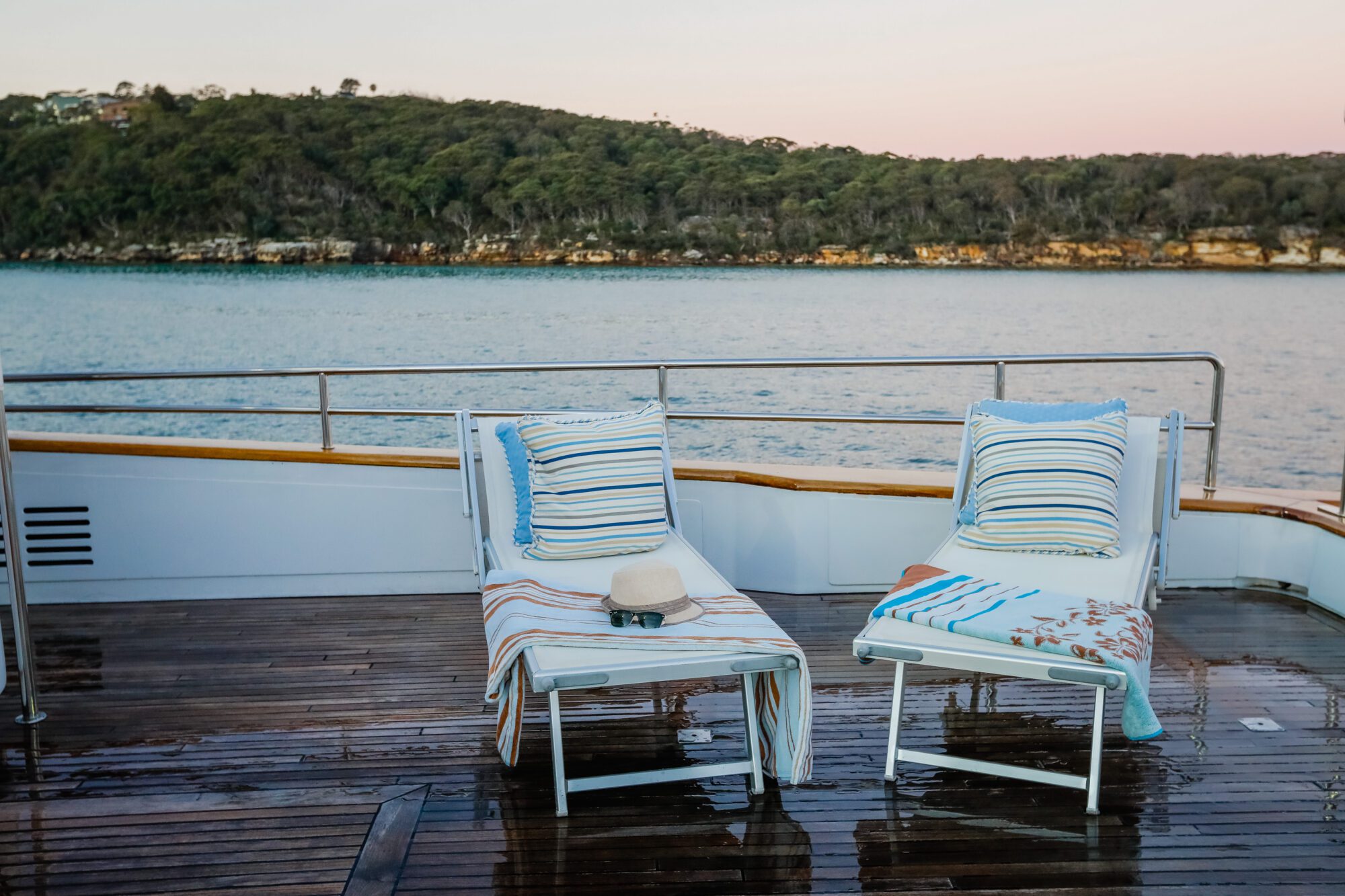 Two lounge chairs with striped cushions and towels are on a wet deck of a boat, facing the water. A hat and sunglasses rest on one chair. An island with trees is visible in the background under a pastel sky.