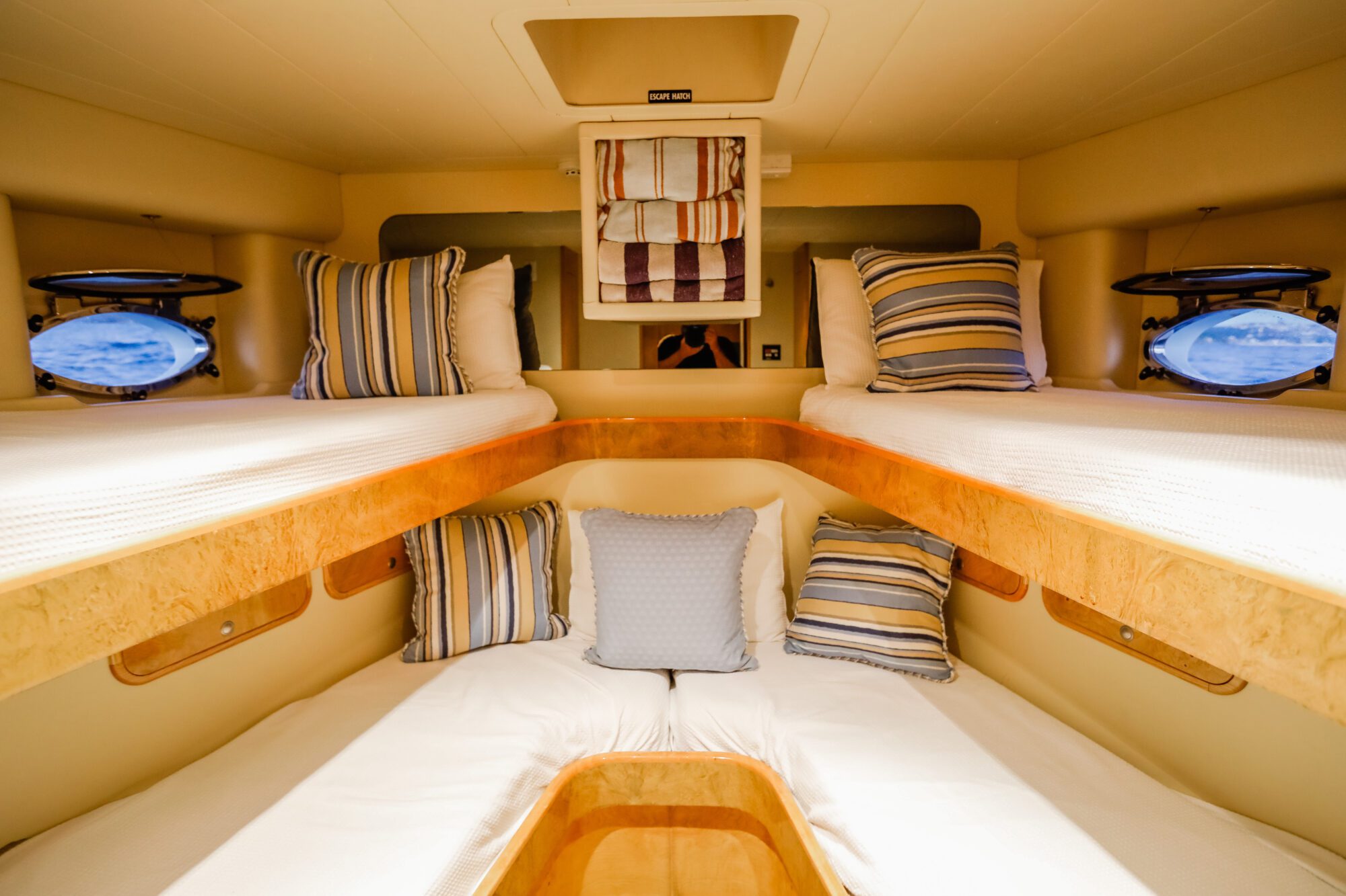 A cozy cabin with four bunk beds, each made up with white linens and striped throw pillows. Small round windows on both sides provide a view of the sea, and warm lighting brightens the space.