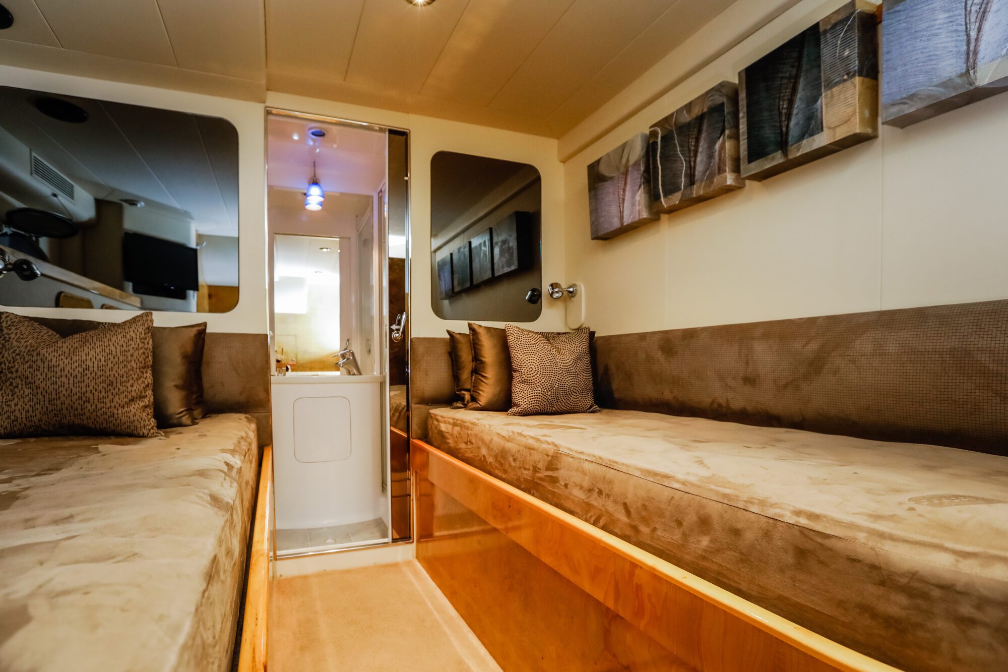 Interior of a boat cabin with two beige cushioned beds, brown patterned pillows, abstract art on the walls, and a small bathroom visible through an open door at the end of the room.