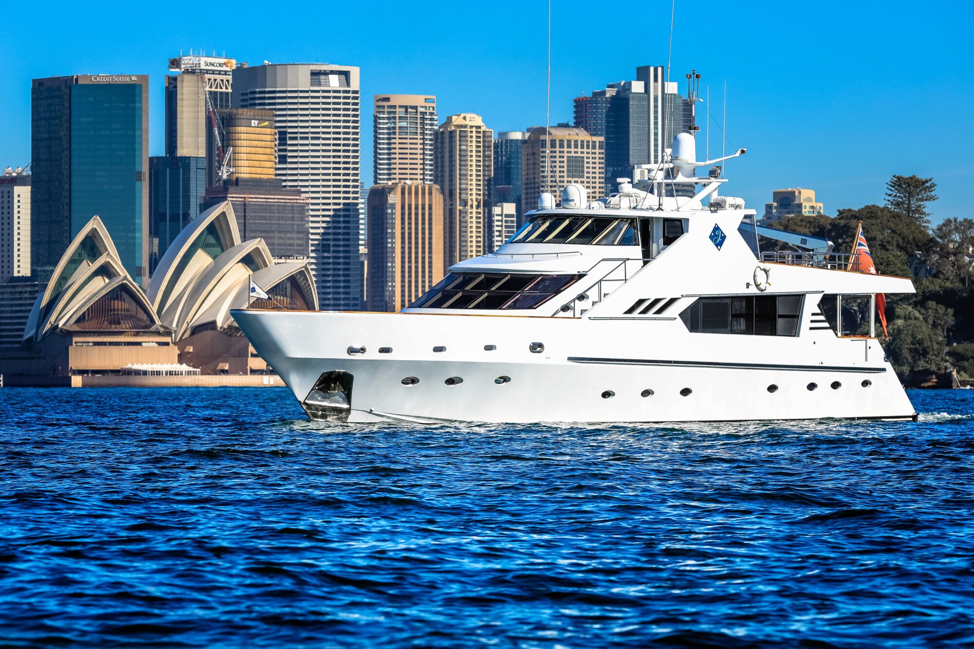 A white luxury yacht sails on blue water with the Sydney Opera House and city skyscrapers in the background under a clear blue sky.