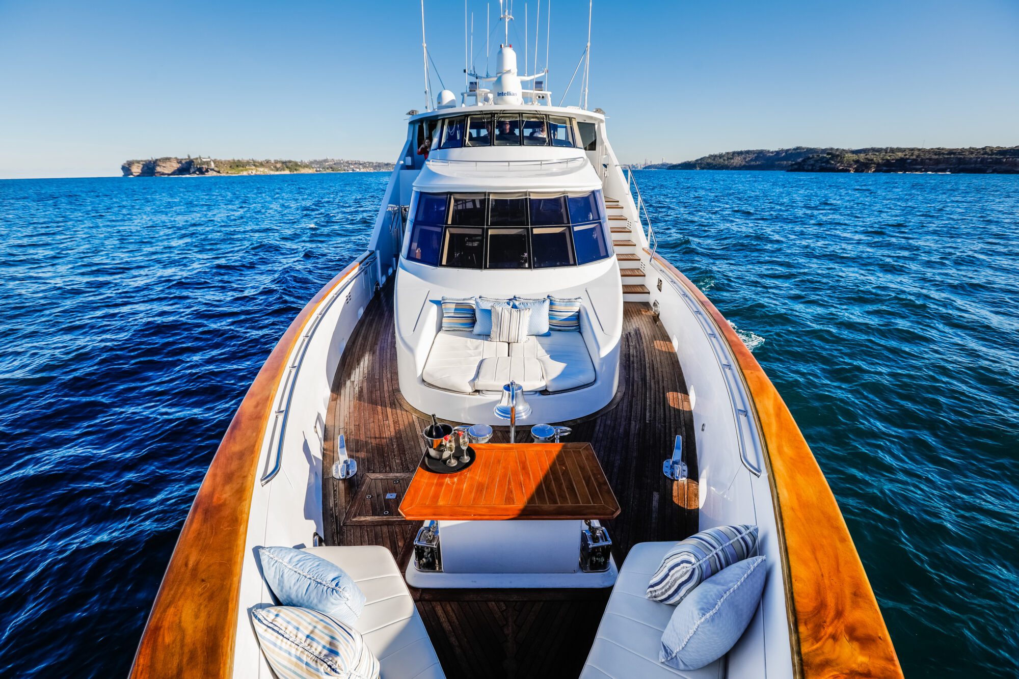 The deck of a luxury yacht with cushioned lounge areas, a wooden table set with drinks, and calm blue ocean water surrounding the boat under a clear sky. Distant land is visible on the horizon.