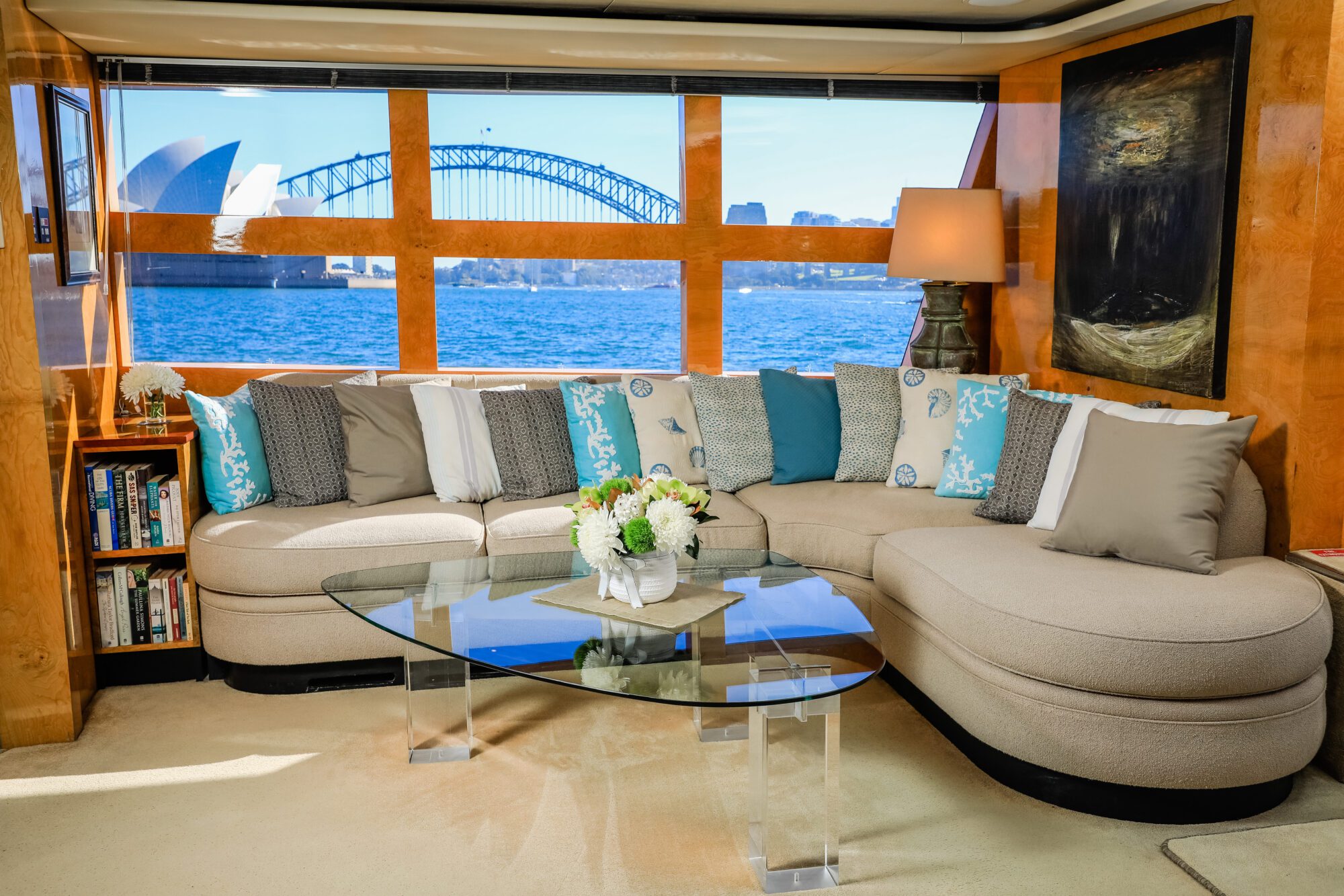 A stylish living room with a beige curved sofa, blue and gray pillows, a glass coffee table with a flower arrangement, and large windows showing the Sydney Opera House and Harbour Bridge in the background.
