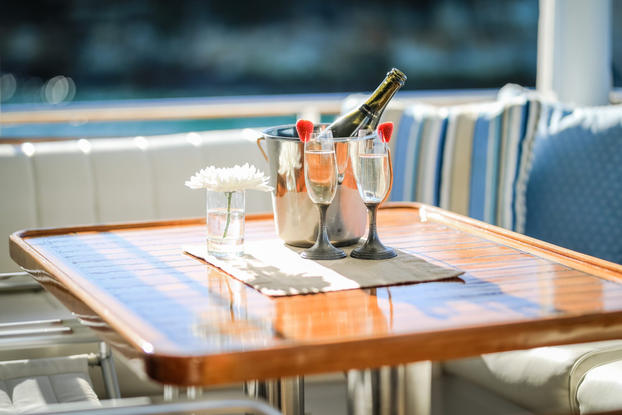 A wooden table set with two glasses of champagne, a bottle in an ice bucket, and a small vase with white flowers, on a sunny patio with blue-striped cushions in the background.
