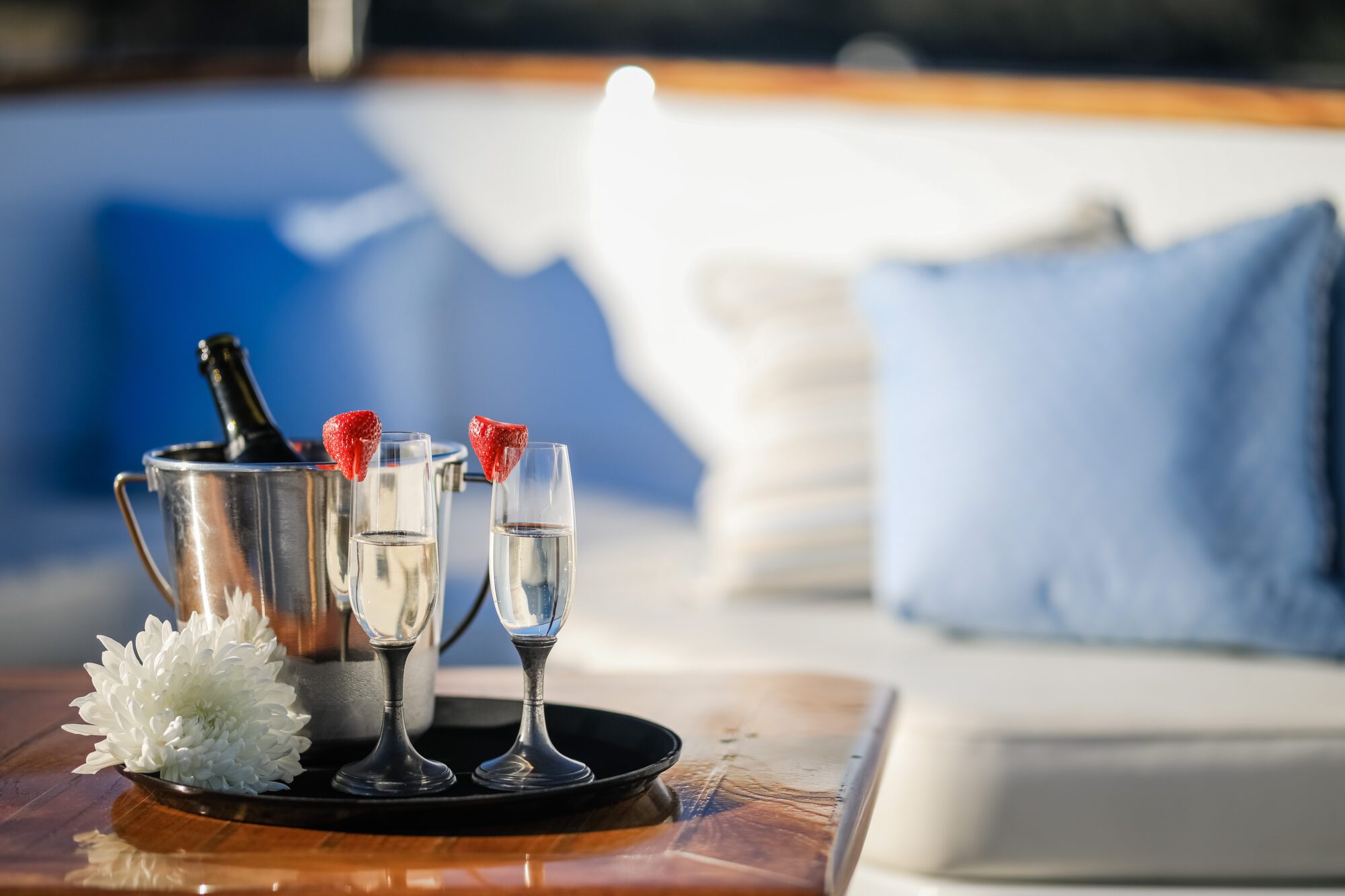 Two champagne flutes with strawberries on the rim and a white flower on a tray, alongside a metal ice bucket with a bottle, set on a wooden table with blue and white pillows in the background.