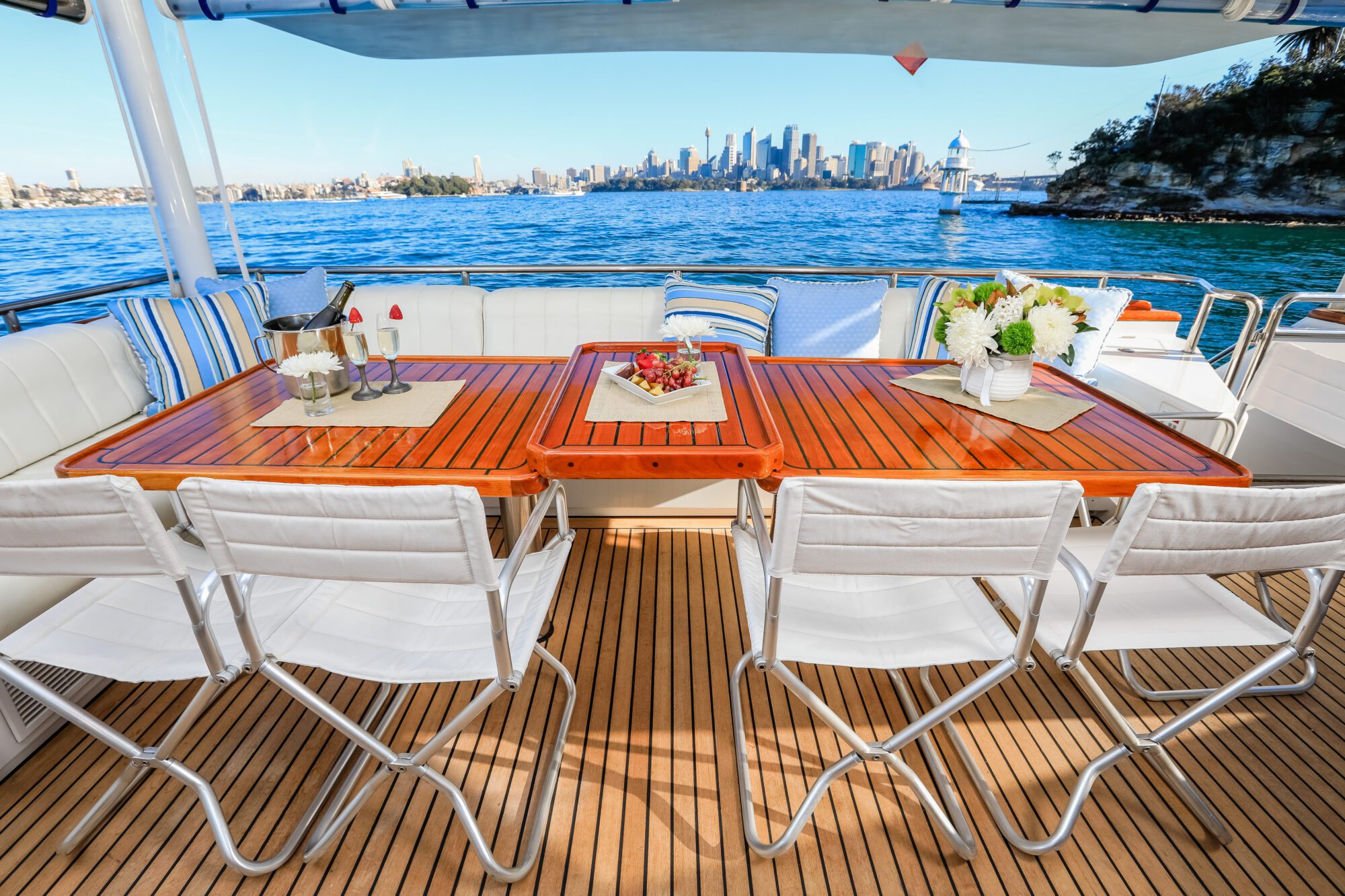 A wooden dining table with white chairs is set on a boat deck, decorated with flowers and fruit, overlooking blue water and a distant city skyline under a clear sky.