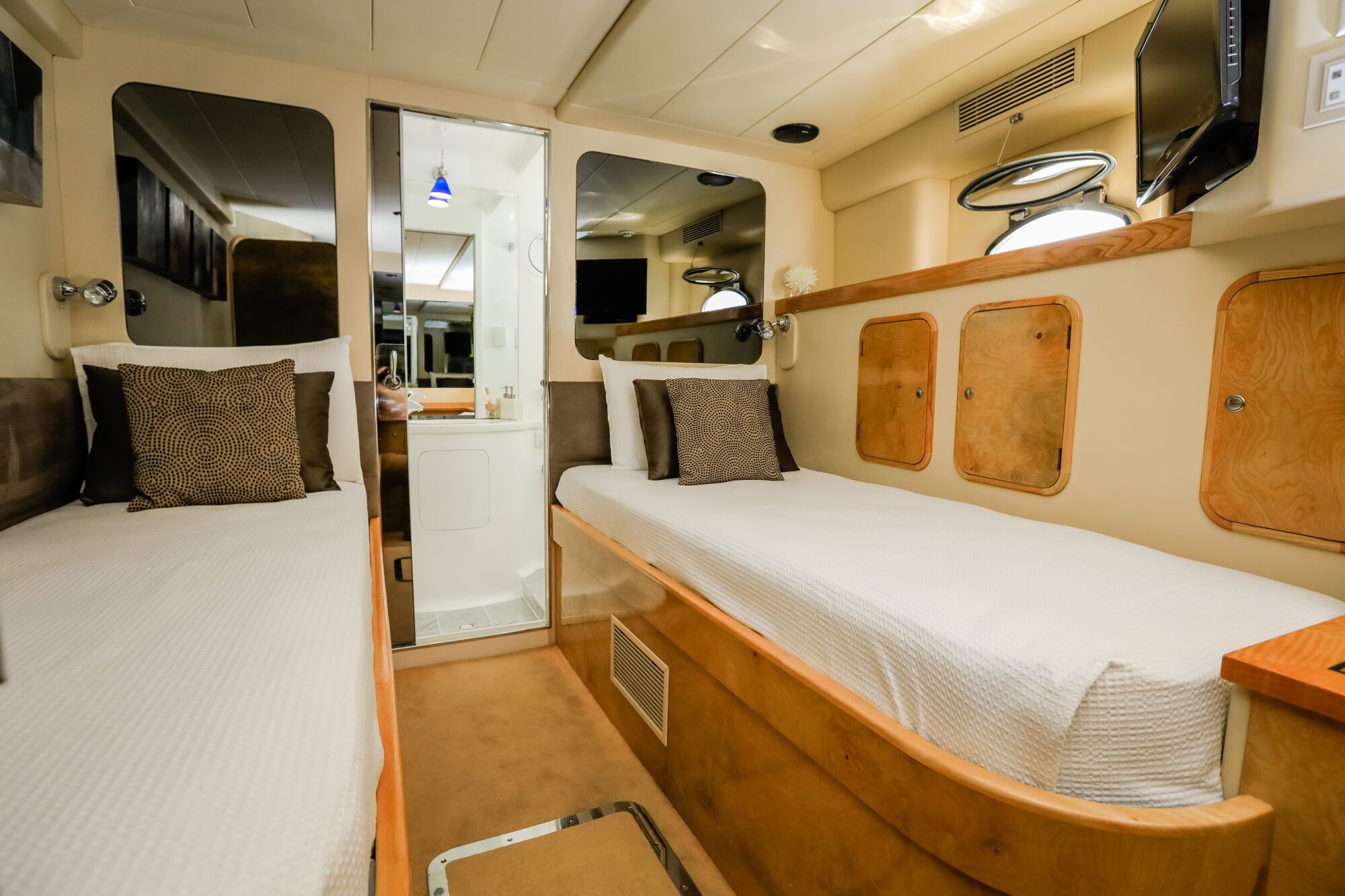 A cozy yacht cabin with two single beds, brown accent pillows, wall-mounted reading lights, small storage cabinets, and a mirror reflecting the bathroom and interior details.