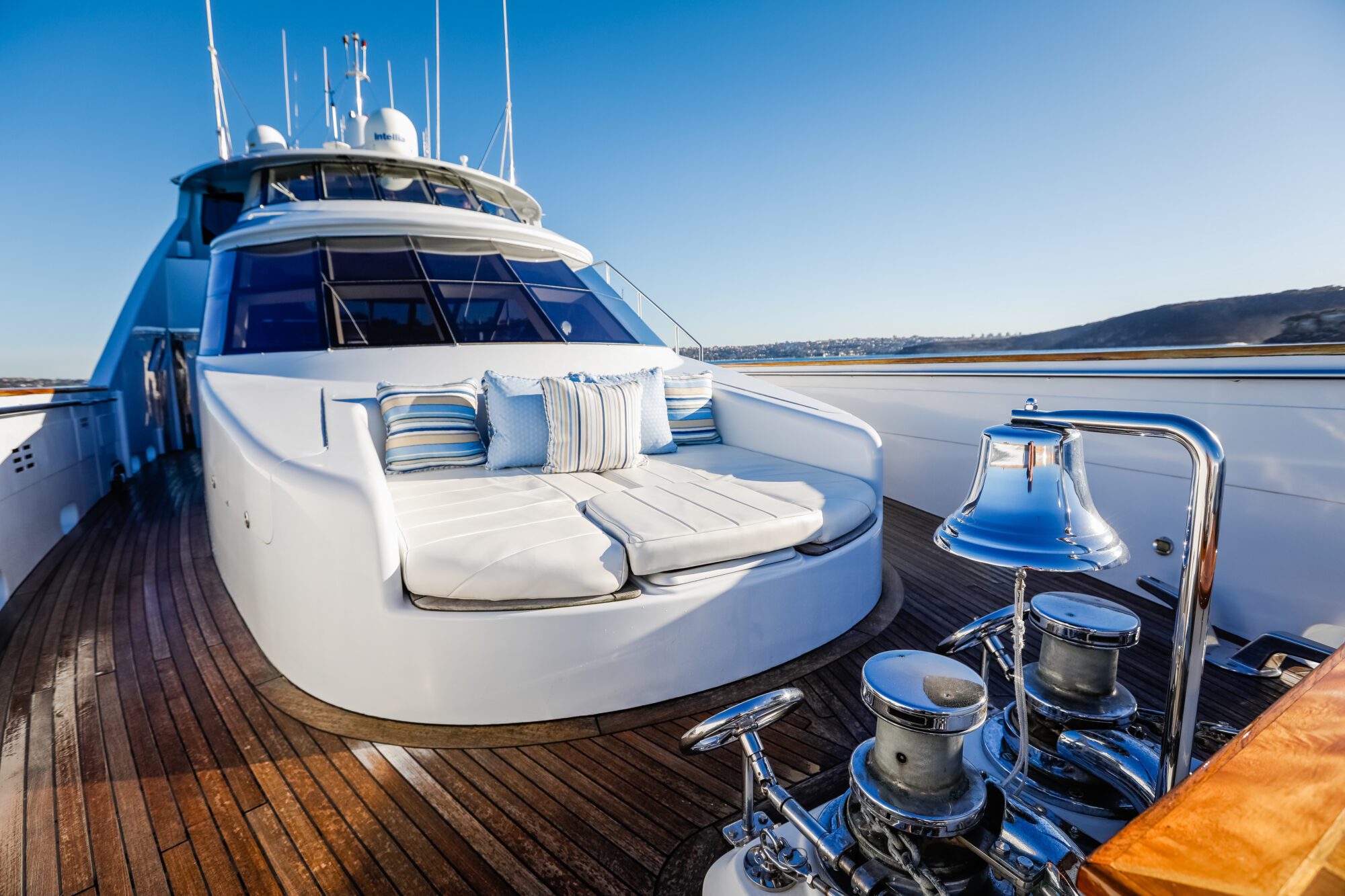 Luxury yacht deck with white cushioned seating, striped pillows, polished wooden flooring, shiny metal fixtures, and a bell, with clear blue sky and distant coastline in the background.
