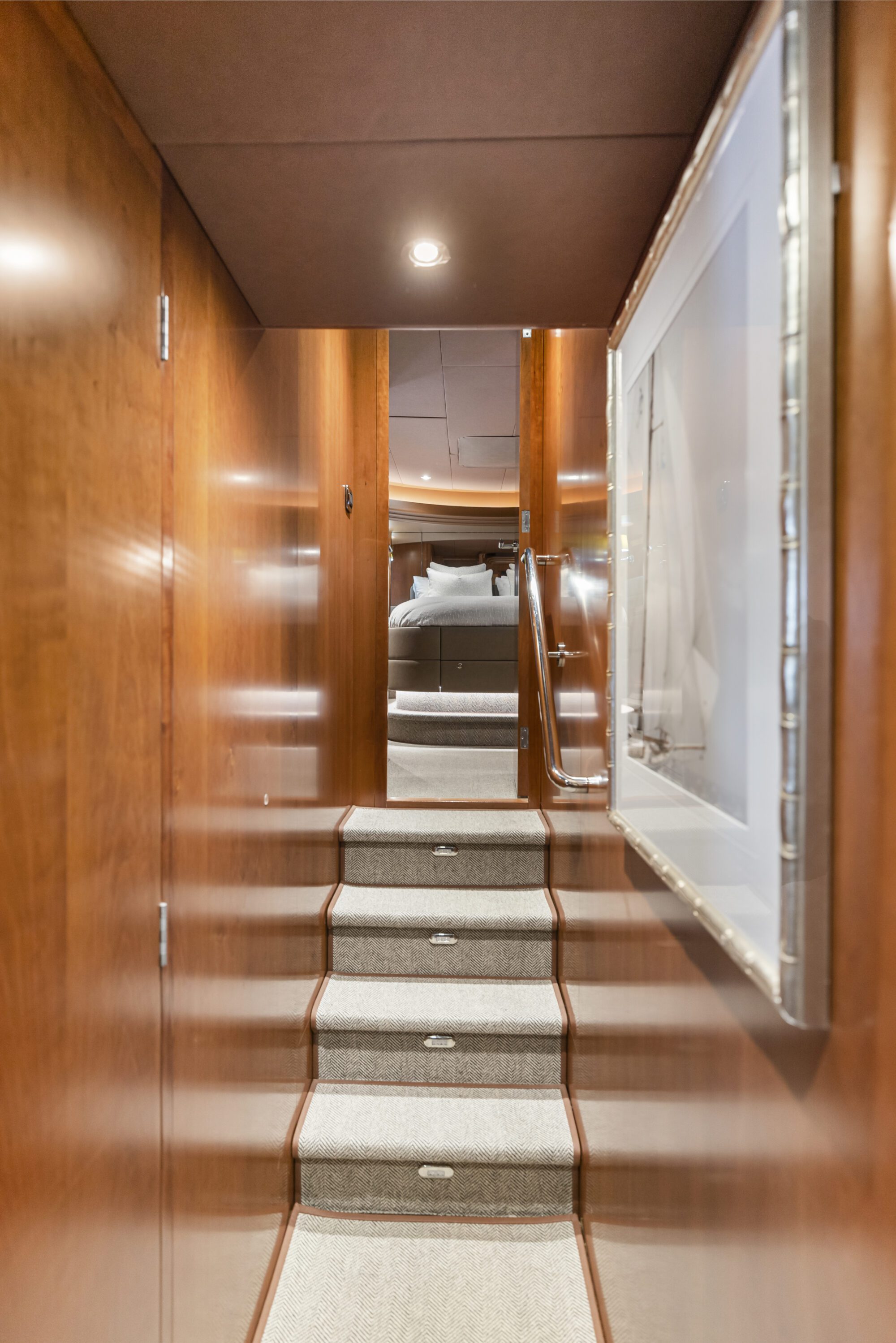 A narrow hallway with wooden walls and a carpeted staircase leading up to a bedroom. A large framed picture hangs on the right wall, and the area is softly lit by a ceiling light.