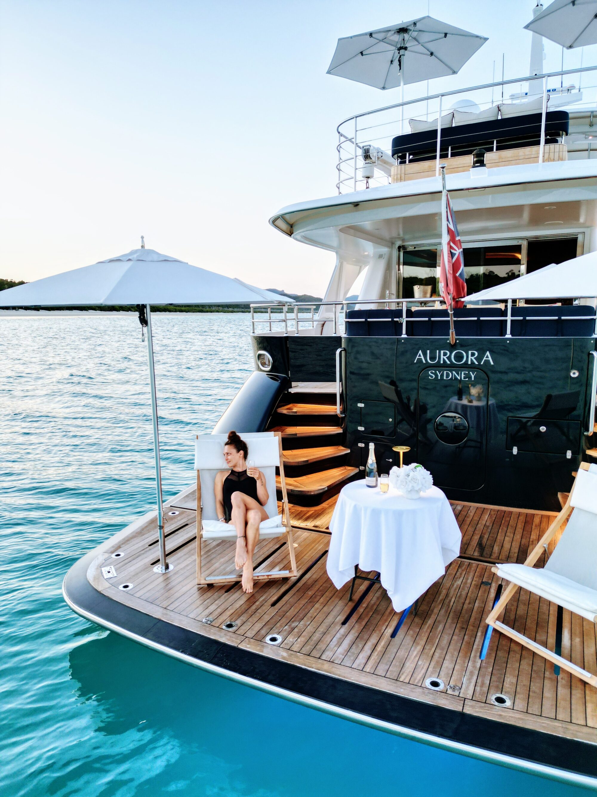 A woman relaxes on a lounge chair on the deck of a luxury yacht named "Aurora Sydney," docked on turquoise water, with a table set for two, parasols, and calm skies in the background.
