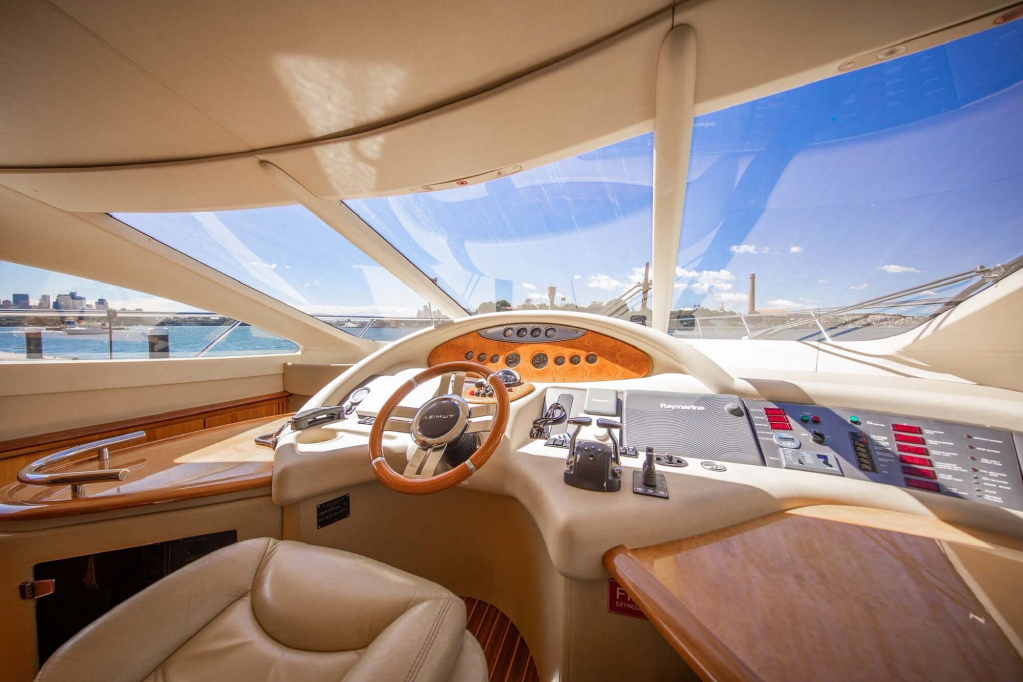 Luxurious yacht cockpit with leather captain’s chair, wooden steering wheel, control panels, and panoramic windows offering a view of a city skyline and blue sky over the water.