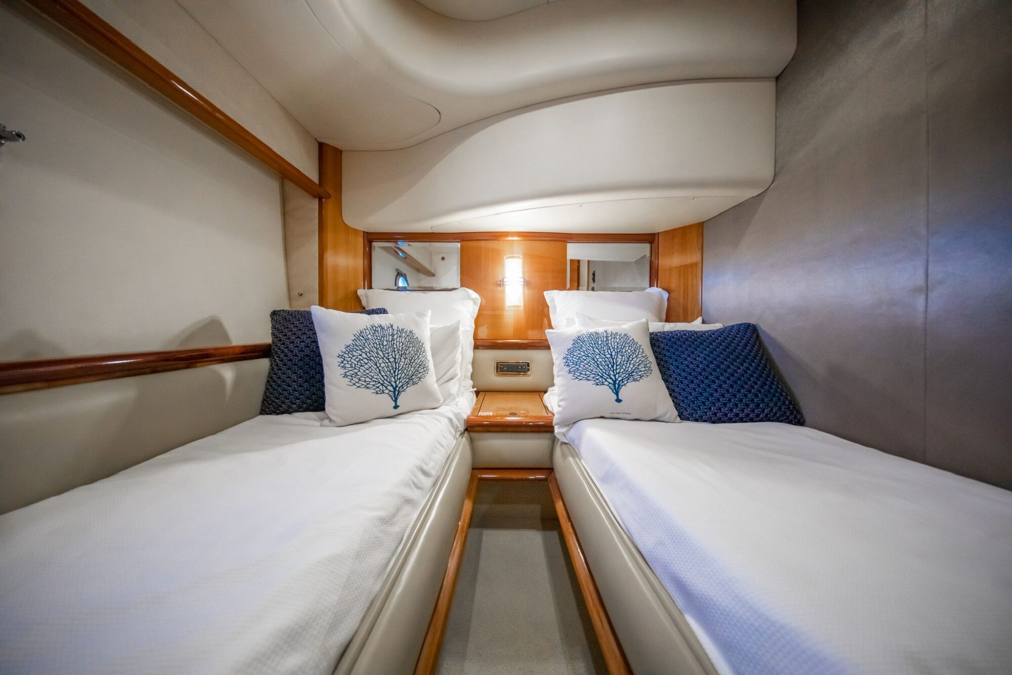A yacht cabin with two single beds, each neatly made with white linens, blue throw pillows, and decorative pillows featuring blue coral designs, separated by a small wooden nightstand.