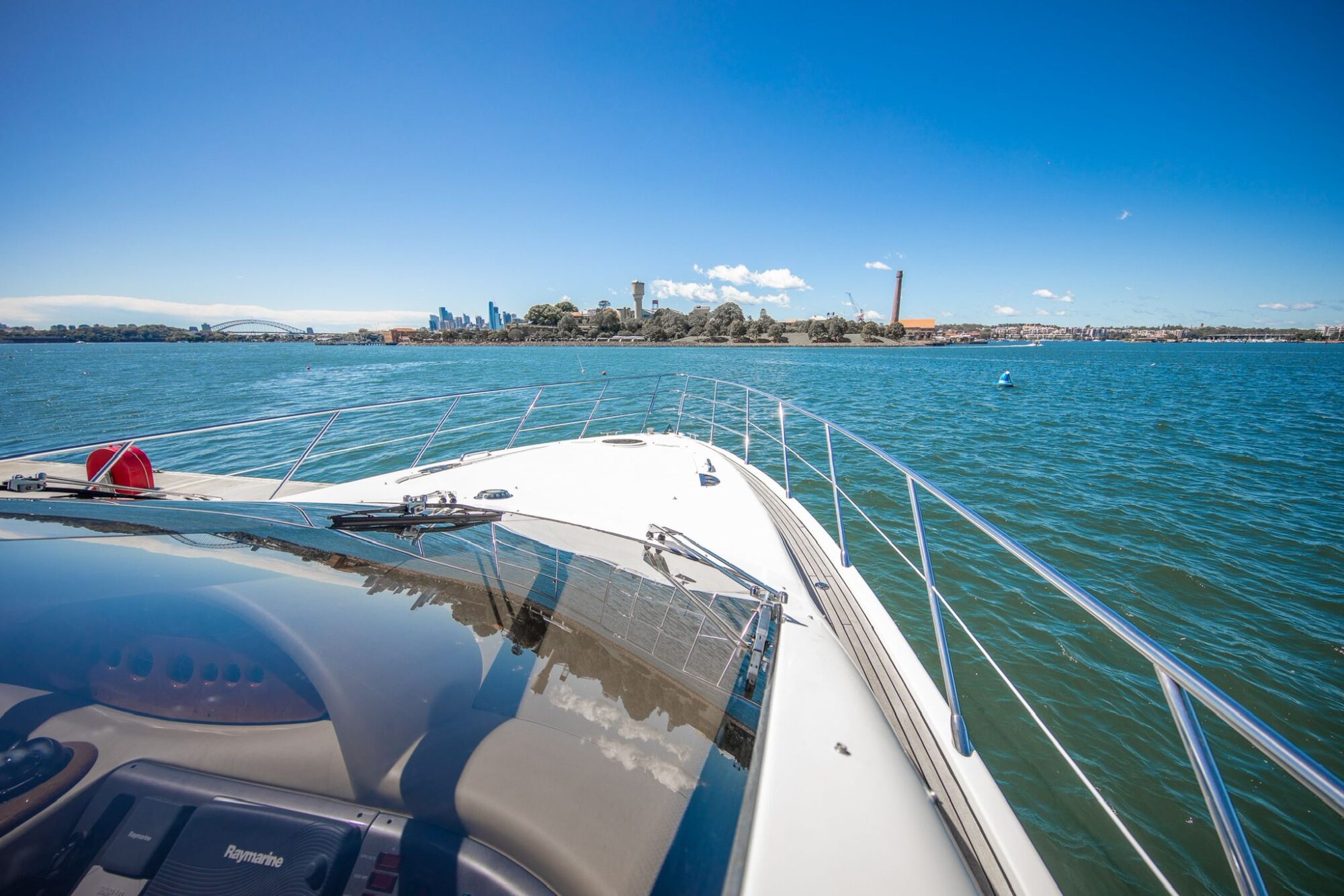 View from the deck of a boat cruising on blue water toward a distant shoreline with trees, buildings, and a city skyline under a bright, clear sky.