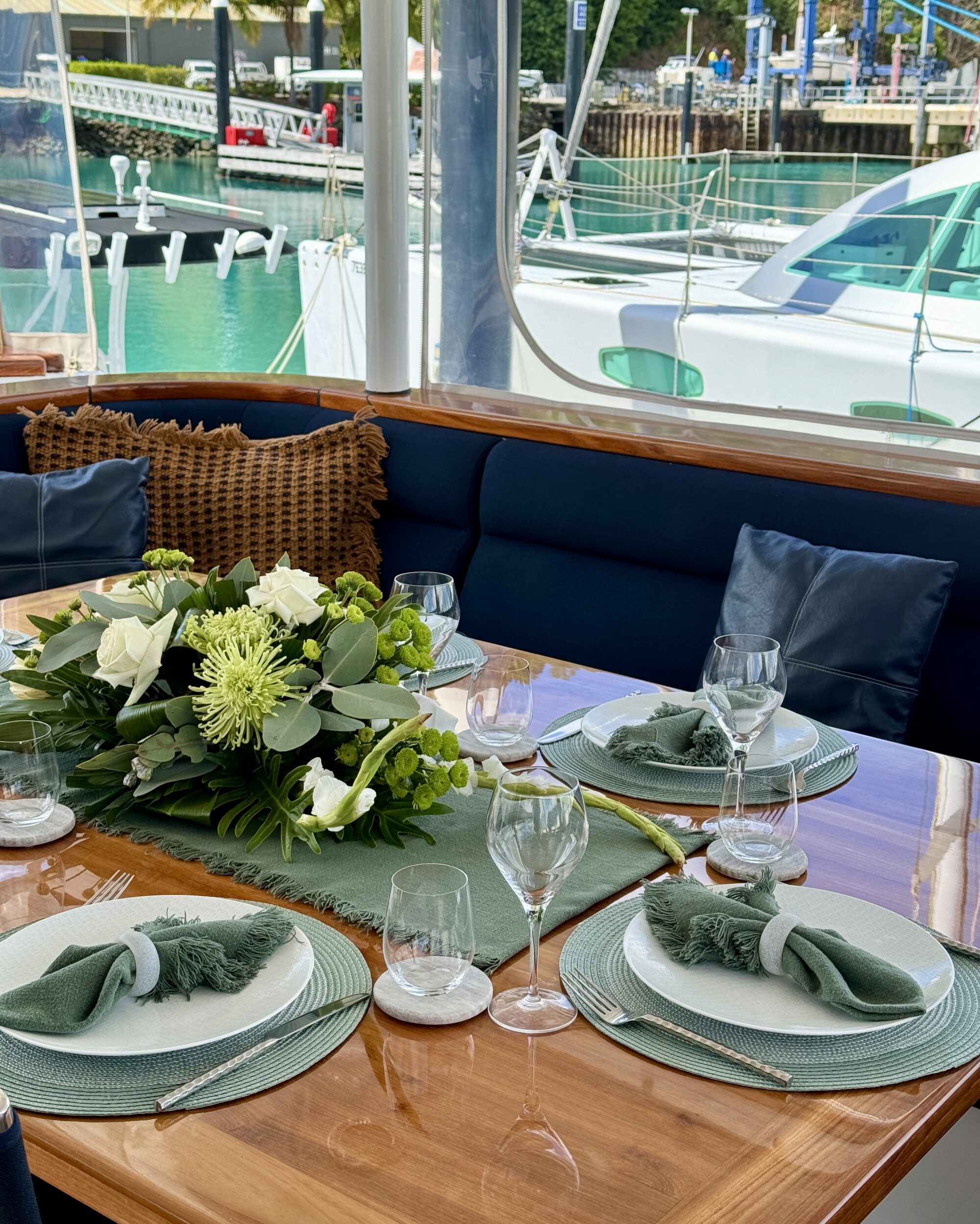 A polished wooden dining table set for four with green napkins, white plates, wine glasses, and a lush green floral centerpiece, on a boat docked at a marina with yachts and turquoise water in the background.