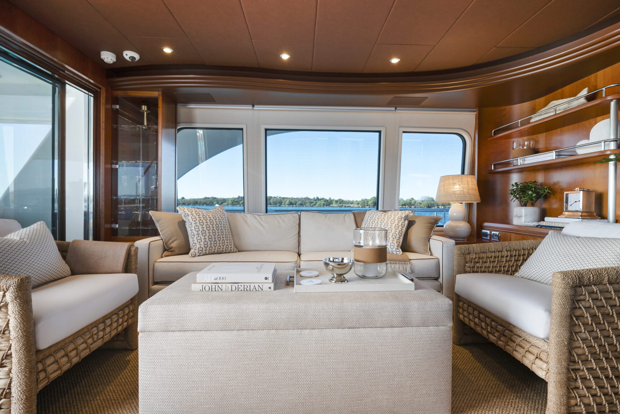 A luxurious yacht lounge with beige sofas, woven armchairs, a cushioned ottoman, and large windows offering a view of the water and trees. Decorative pillows, books, and a lamp add to the cozy, elegant atmosphere.