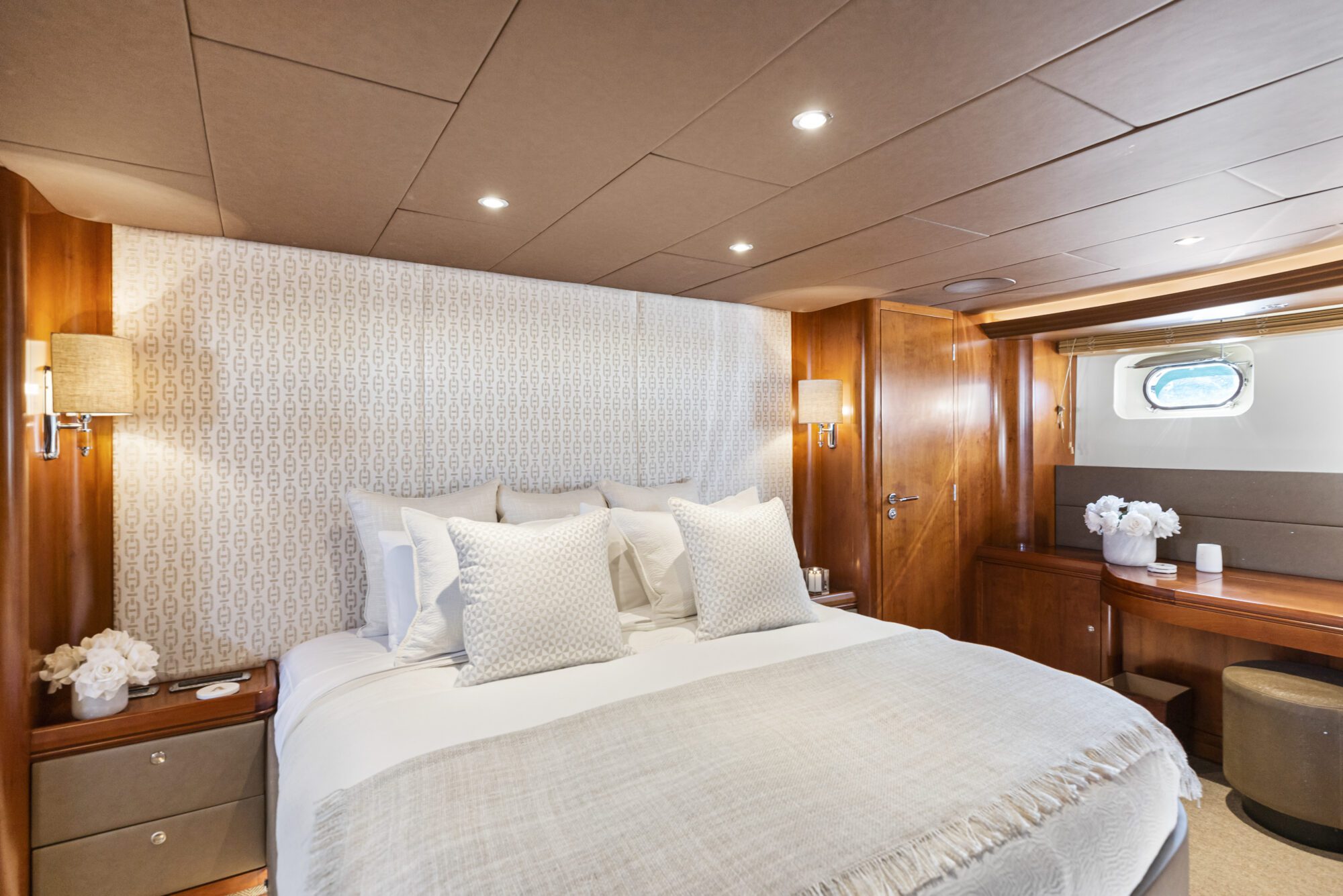 A luxurious yacht bedroom with a large bed, white and beige bedding, patterned headboard, wooden paneling, two lamps, a window, and a vanity with flowers and a mirror.
