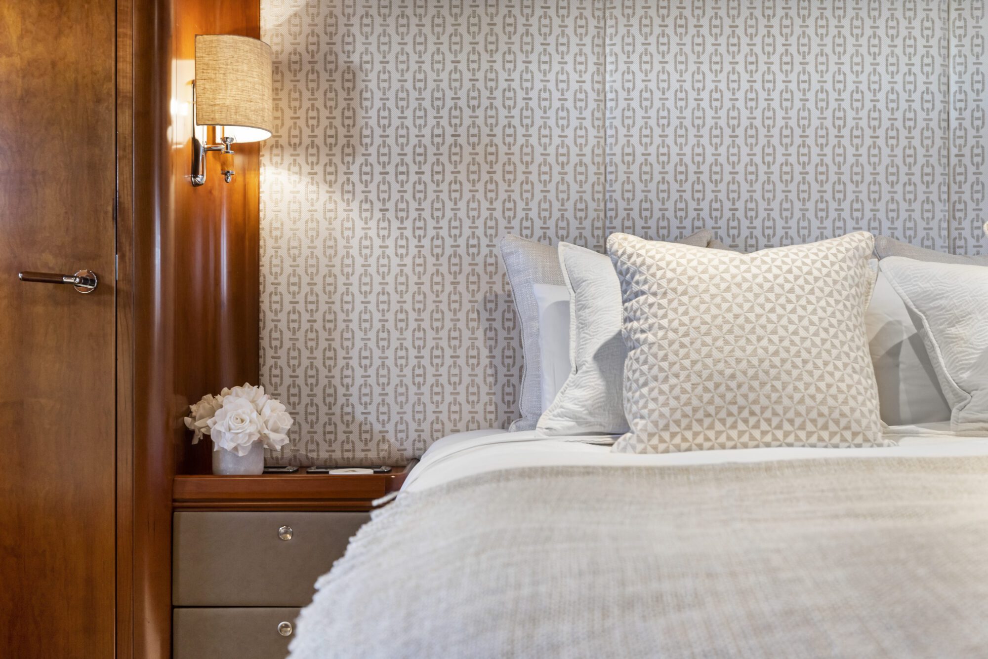 A cozy bedroom with a bed featuring white and textured pillows, a beige patterned wall, a wooden nightstand with white flowers, and a wall sconce with a lighted lamp.