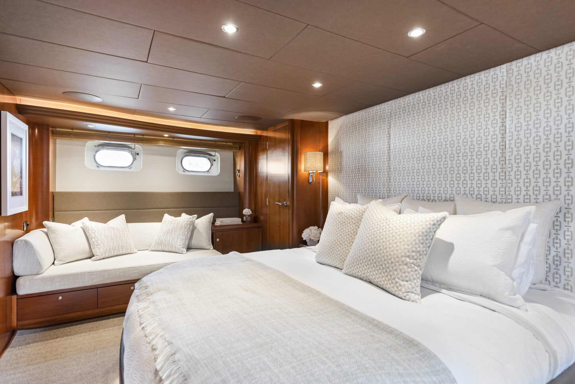 A luxurious yacht bedroom with a large bed, plush pillows, a cozy bench by two round windows, wood paneling, and soft lighting creating a warm, inviting atmosphere.