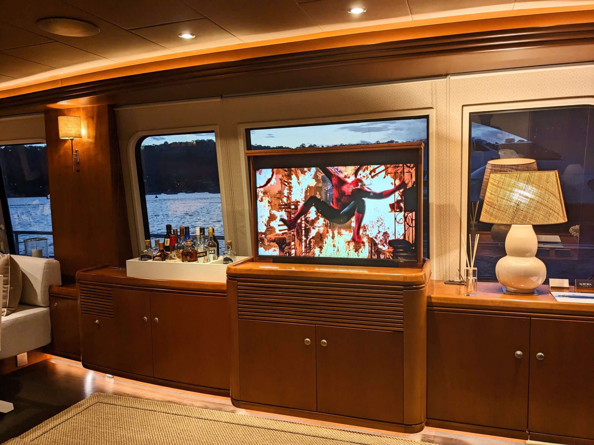 A luxury yacht interior with a wooden cabinet, bottles of liquor, a lamp, and a TV displaying a colorful action scene. Large windows show water and a scenic view outside at sunset.