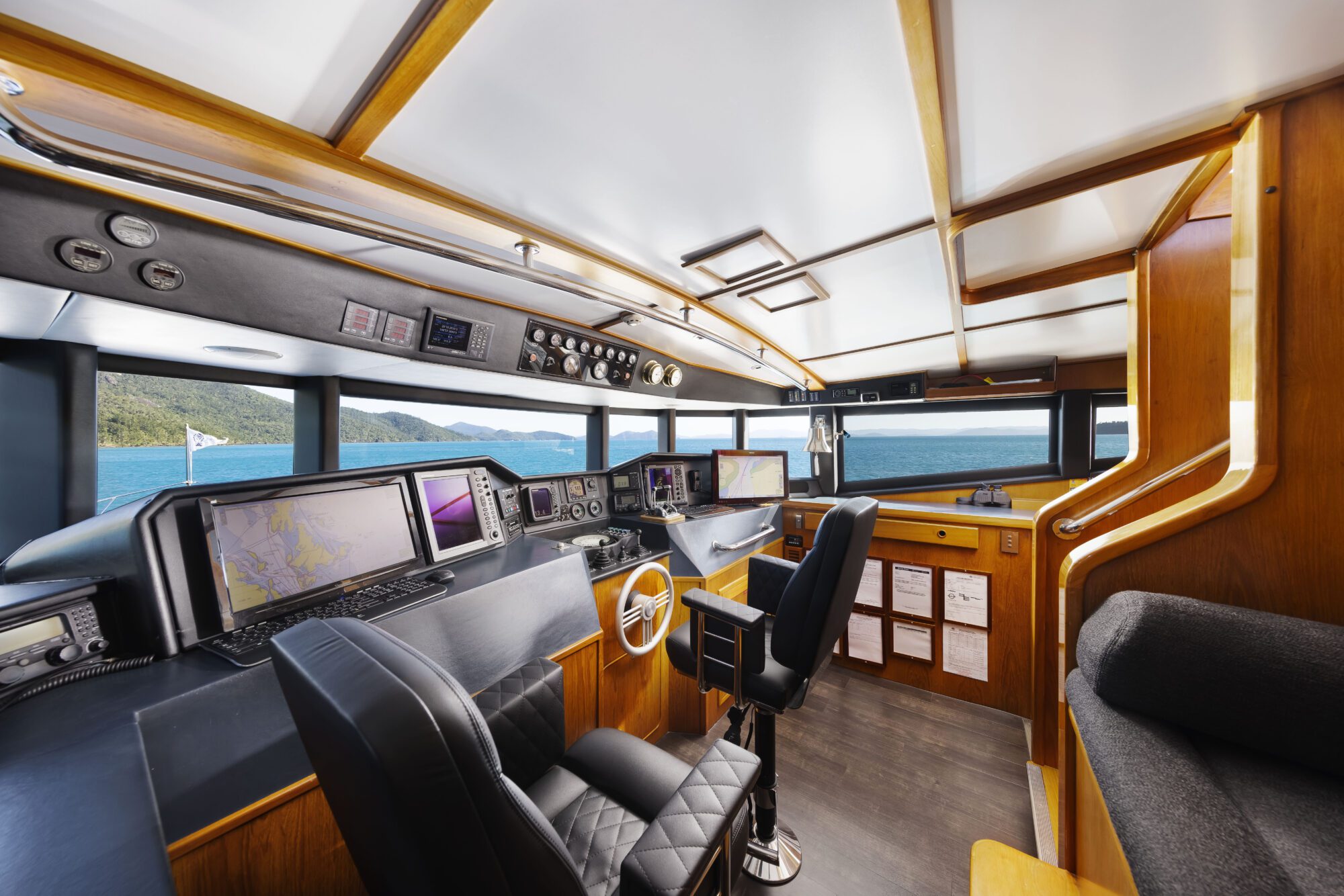 Modern boat cockpit with large windows showing ocean and distant hills. The interior has wooden panels, navigation screens, controls, black padded chairs, and a staircase on the right. Bright natural light fills the space.
