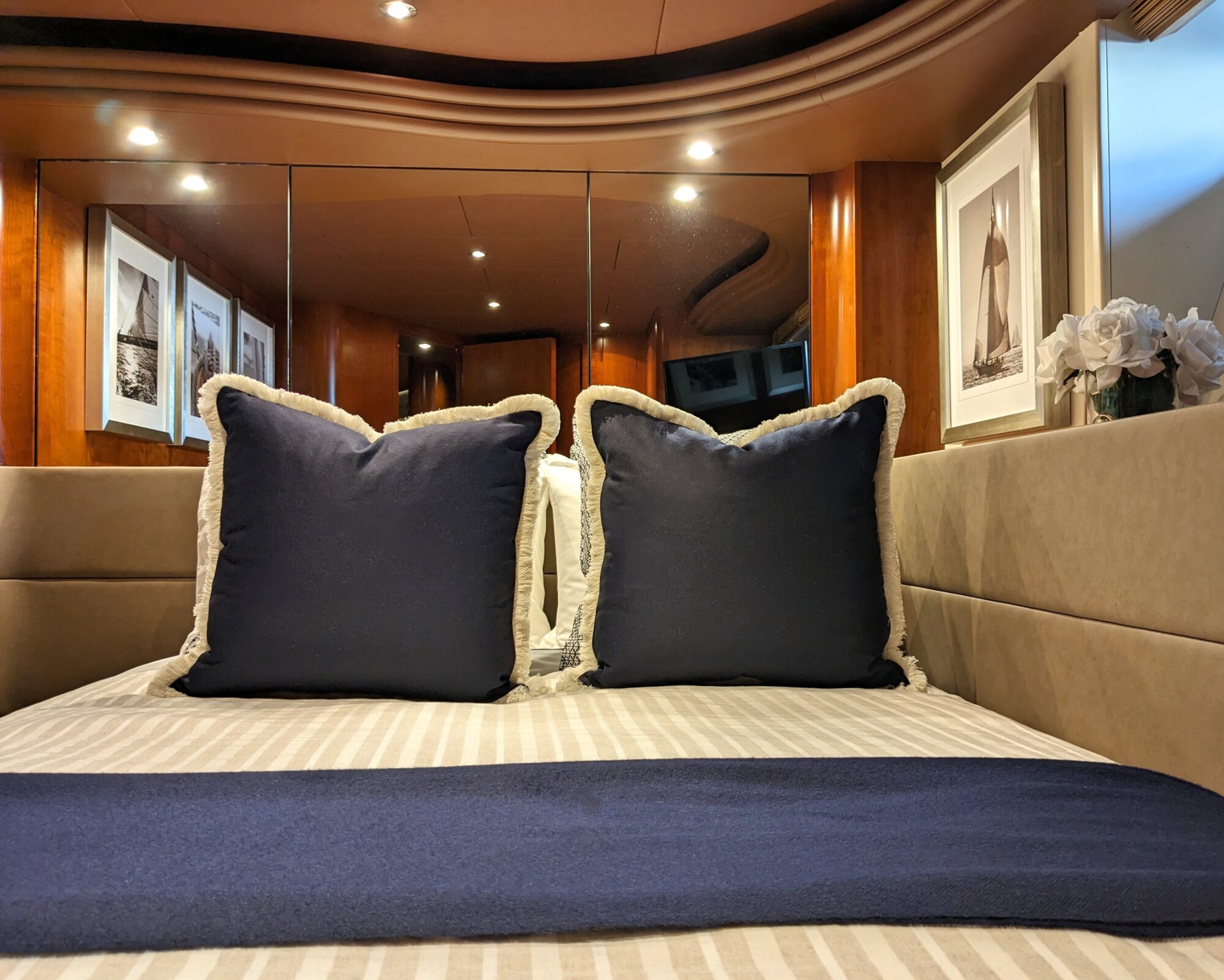 A neatly made bed with two navy blue pillows, a matching throw, beige striped bedding, and framed nautical artwork on wood-paneled walls, with mirrors reflecting the cozy, well-lit bedroom.