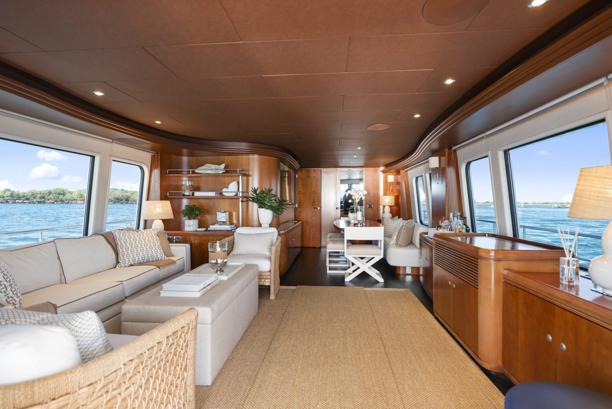 Spacious yacht interior with large windows showing water views, featuring light-colored sofas, wooden paneling, a woven rug, built-in shelves, and soft lighting creating a cozy, luxurious atmosphere.