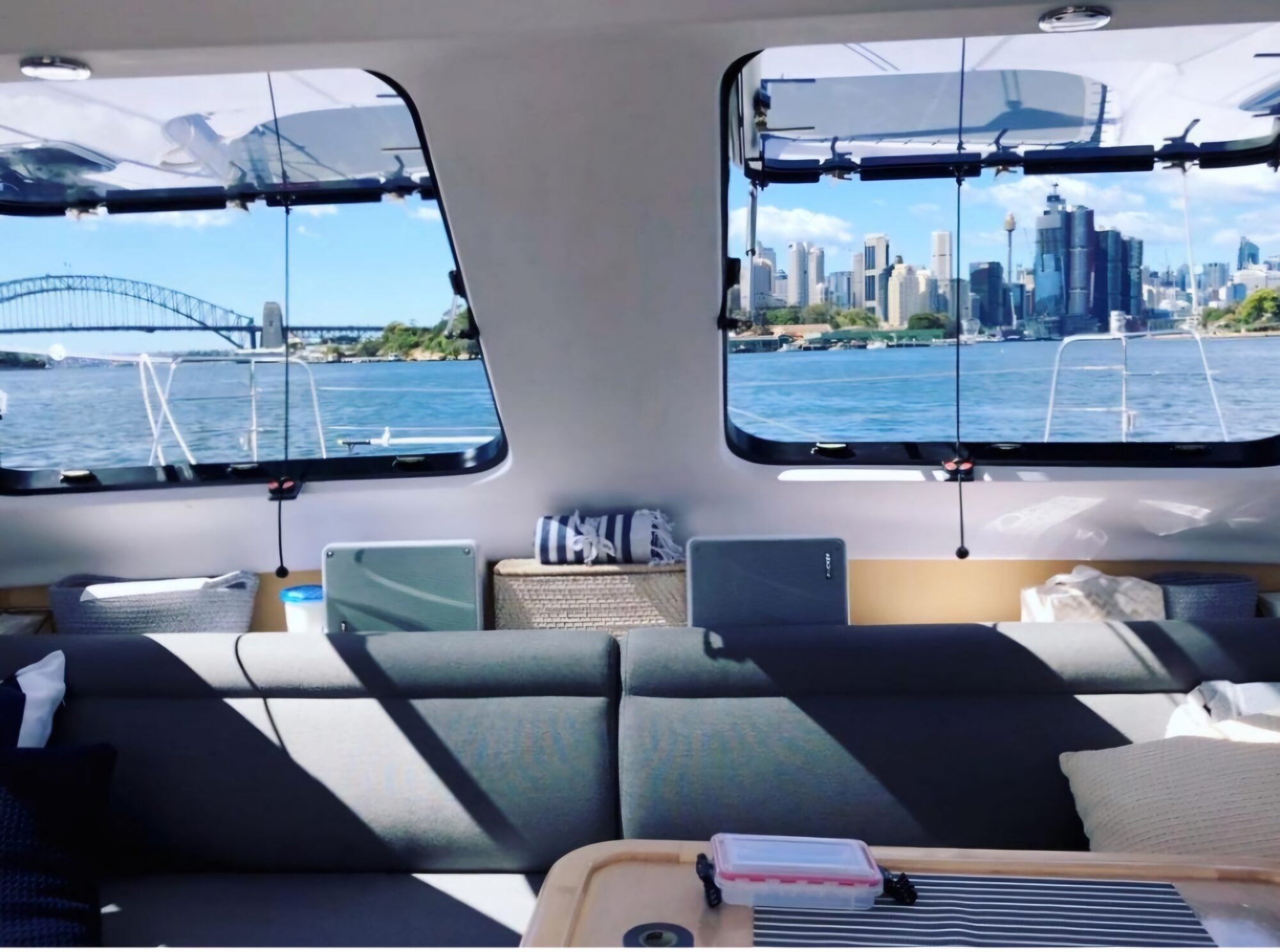 A view of Sydney’s skyline and Harbour Bridge through the windows of a boat, with sunlight illuminating the modern interior seating area and table in the foreground.