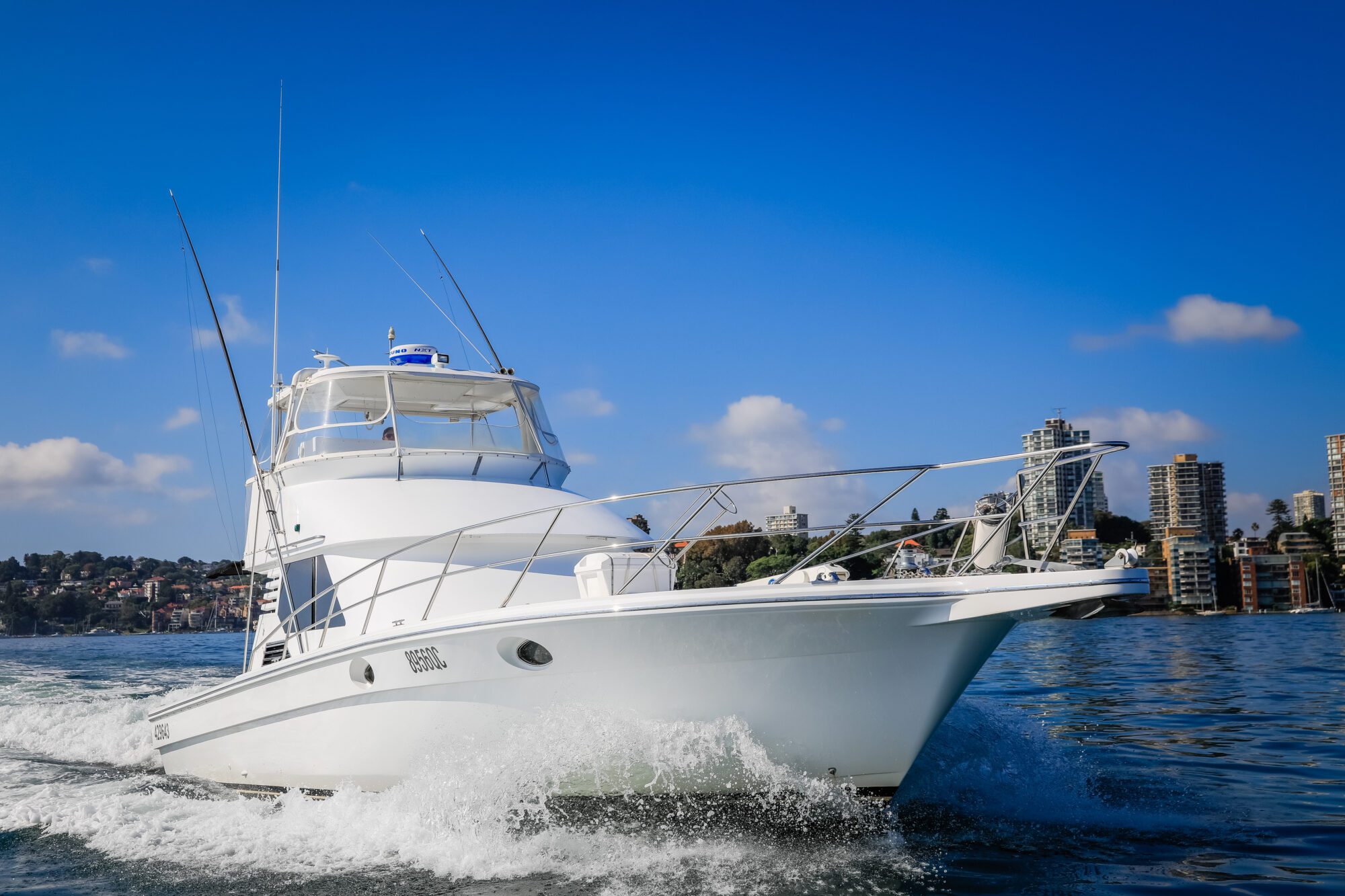 A white motor yacht cruises through calm water with a city skyline and trees in the background under a clear blue sky.