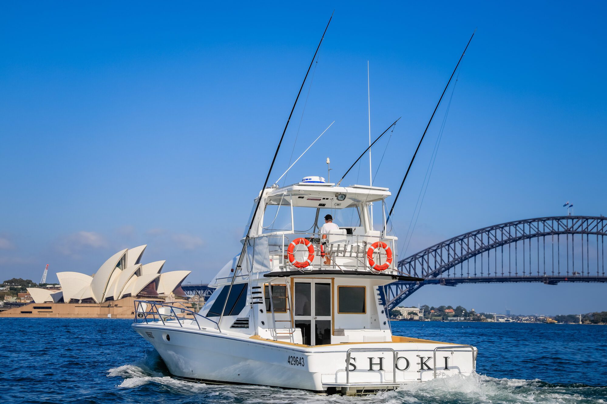 A white boat named "SHOKI" sails on Sydney Harbour, with the Sydney Opera House and Harbour Bridge in the background under a clear blue sky.