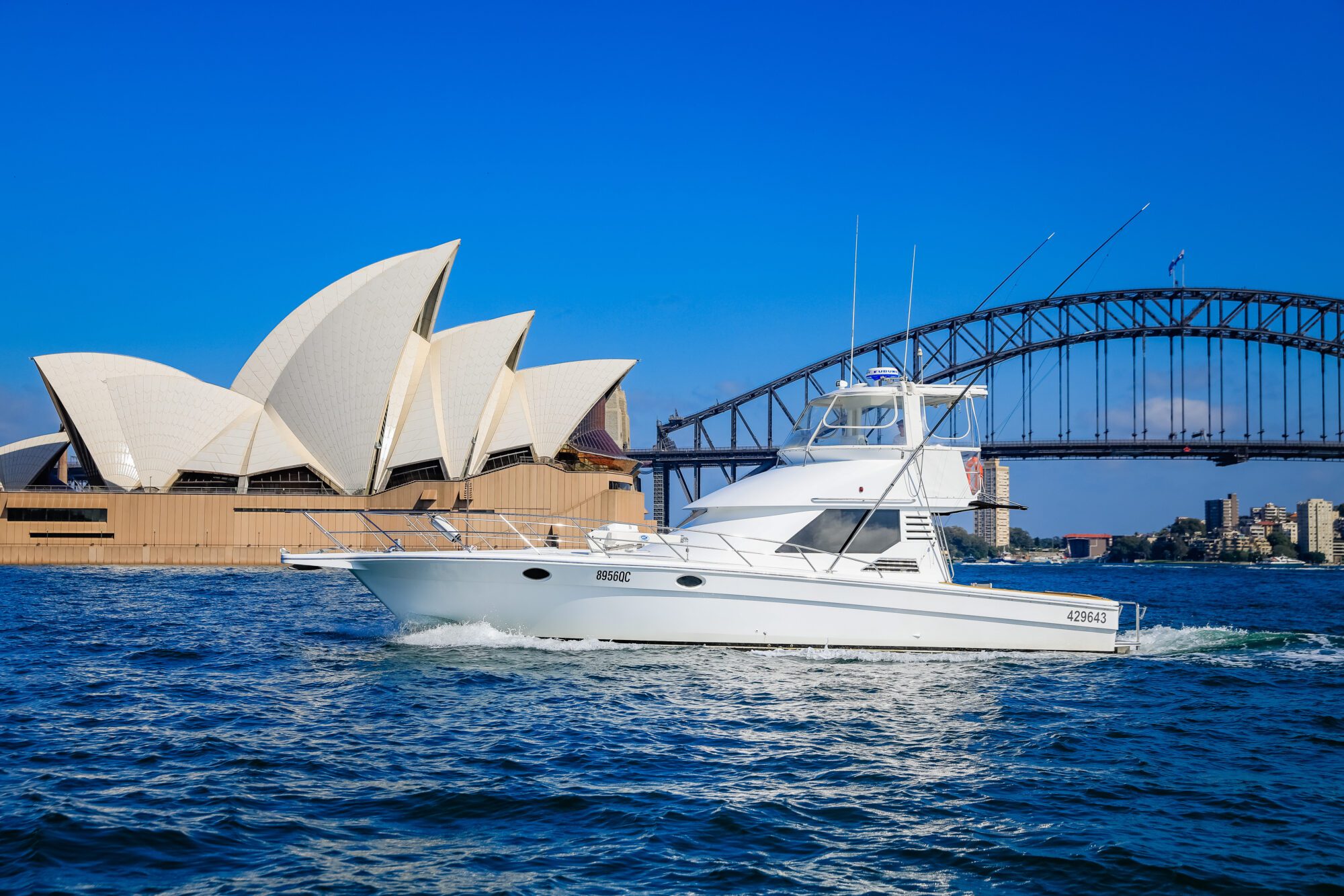 A white motor yacht cruises on Sydney Harbour with the Sydney Opera House and Harbour Bridge in the background under a clear blue sky.