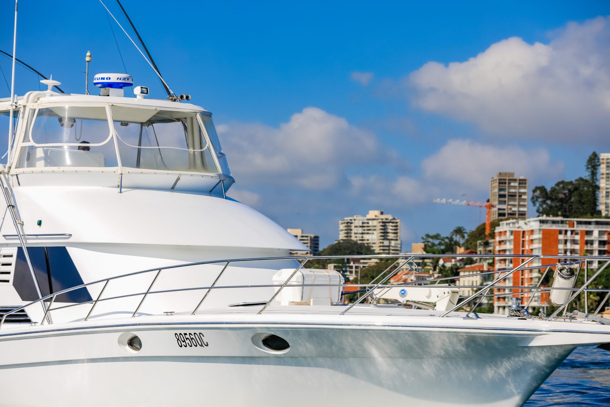 A white yacht is docked in the water with tall city buildings and a crane visible in the background under a partly cloudy blue sky.