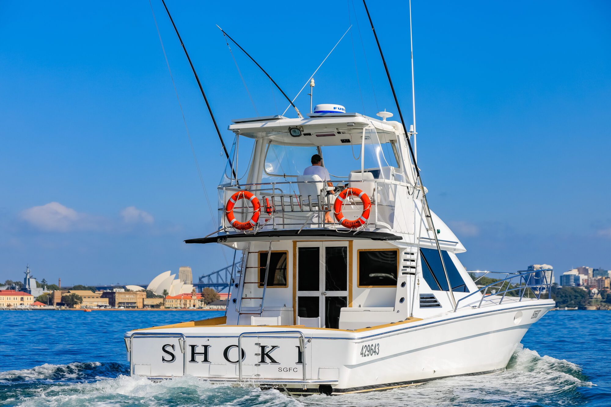 A white motorboat named "SHOK" cruises through blue water near a city skyline, with a person at the helm and two orange life rings attached to the back. The Sydney Opera House and Harbour Bridge are visible in the background.