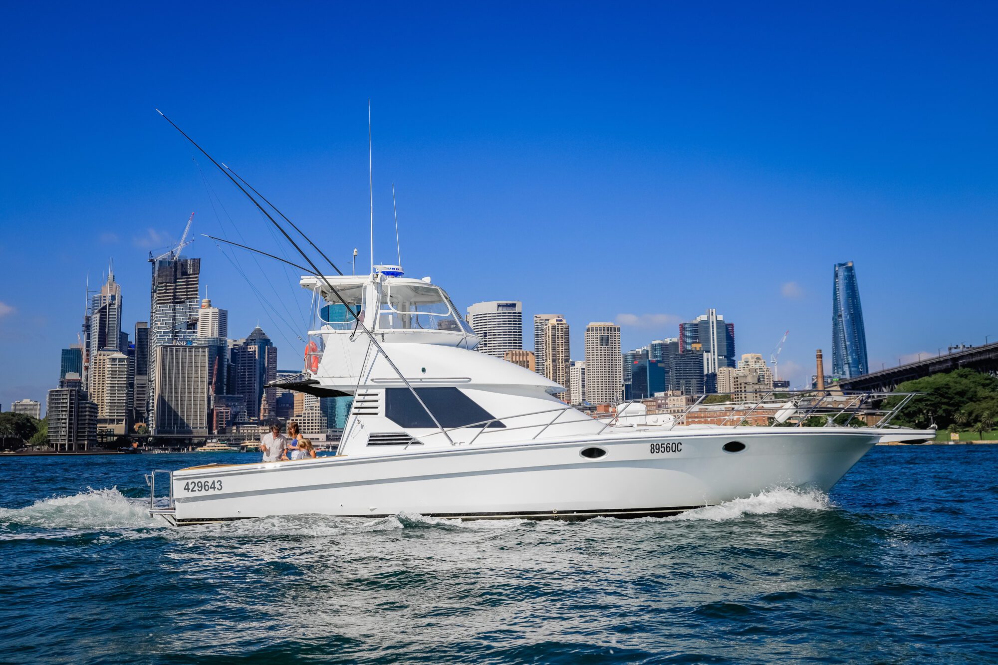 A white motor yacht cruises on blue water, with fishing rods mounted on the back. A modern city skyline with tall buildings and a bridge is visible in the background under a clear blue sky.
