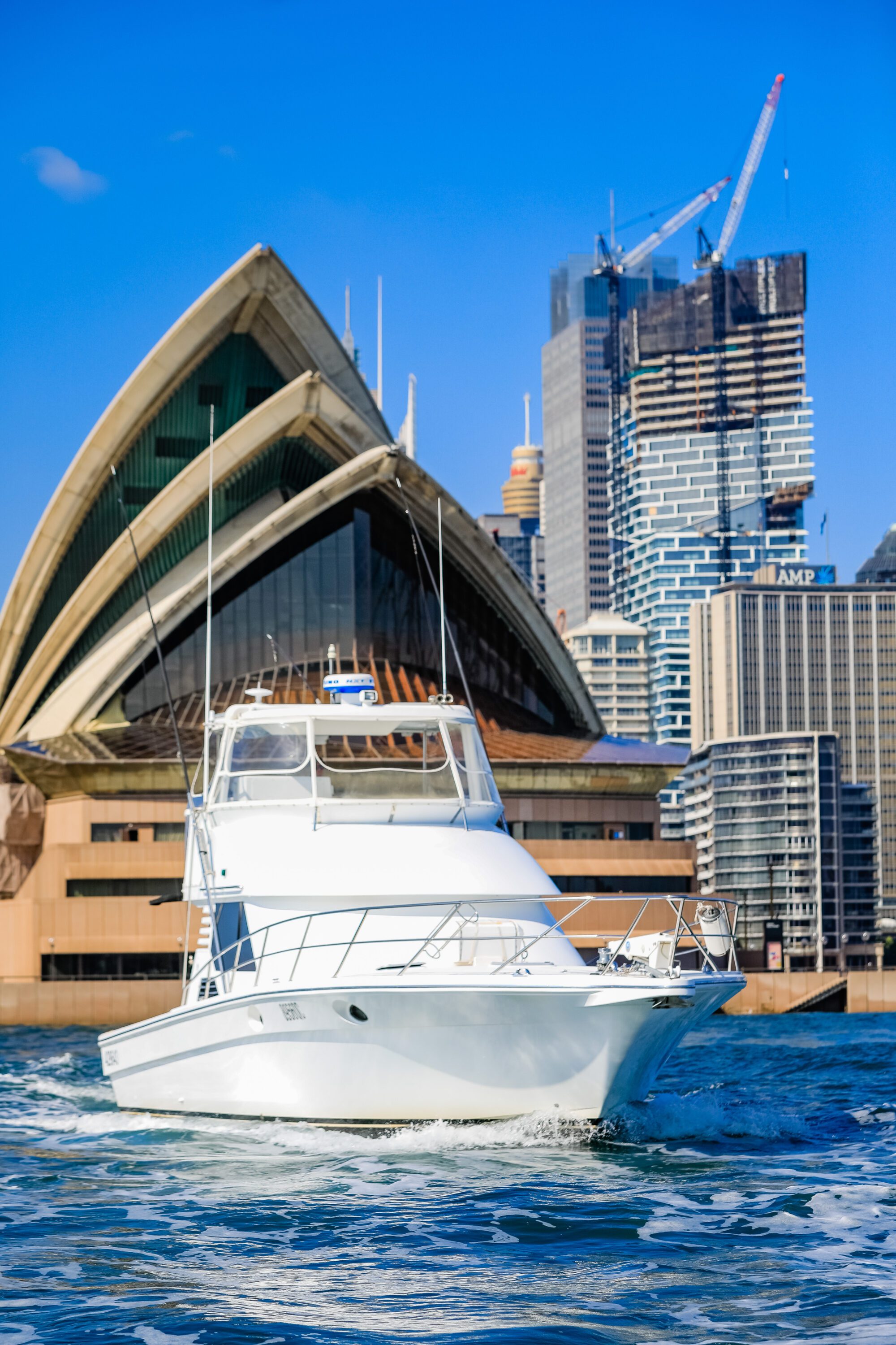 A white yacht cruises on blue water in front of the Sydney Opera House, with modern high-rise buildings and construction cranes in the background under a clear blue sky.