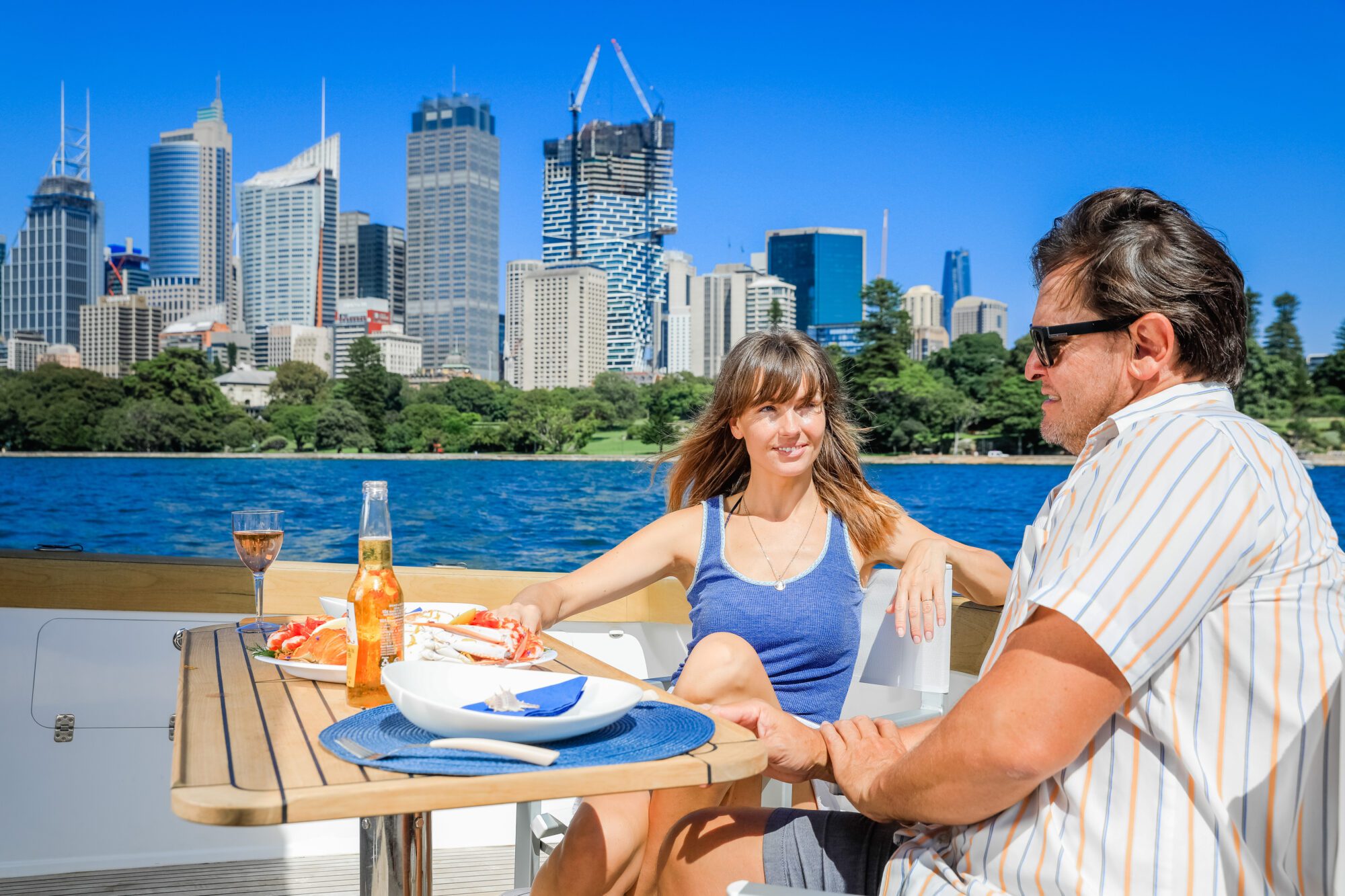 A woman and man relax on a boat, enjoying drinks and seafood at a table. Tall city buildings and clear blue sky are visible in the background across the water.