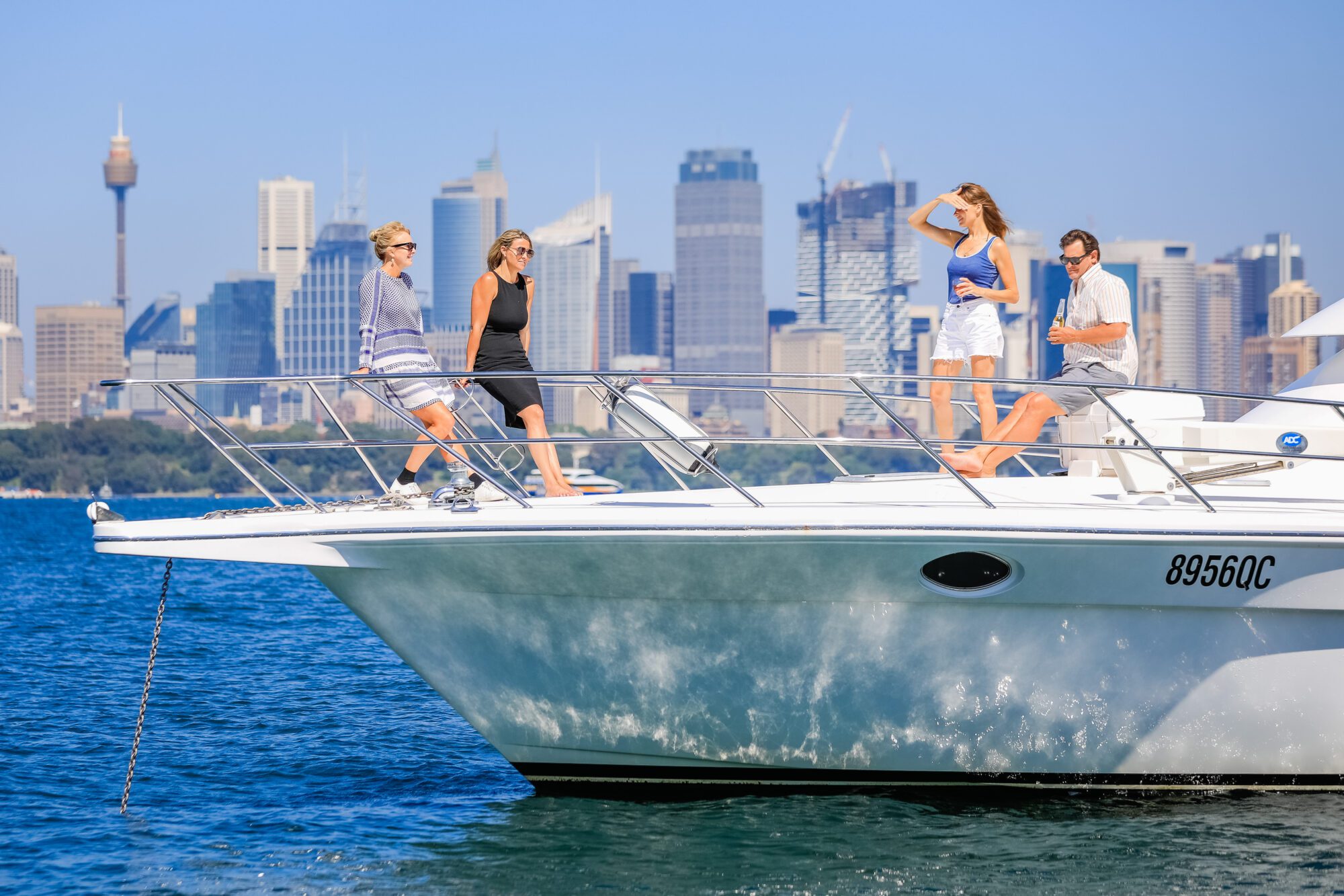 Four people stand and walk on the deck of a white yacht on the water, with a city skyline and tall buildings visible in the background under a clear blue sky.