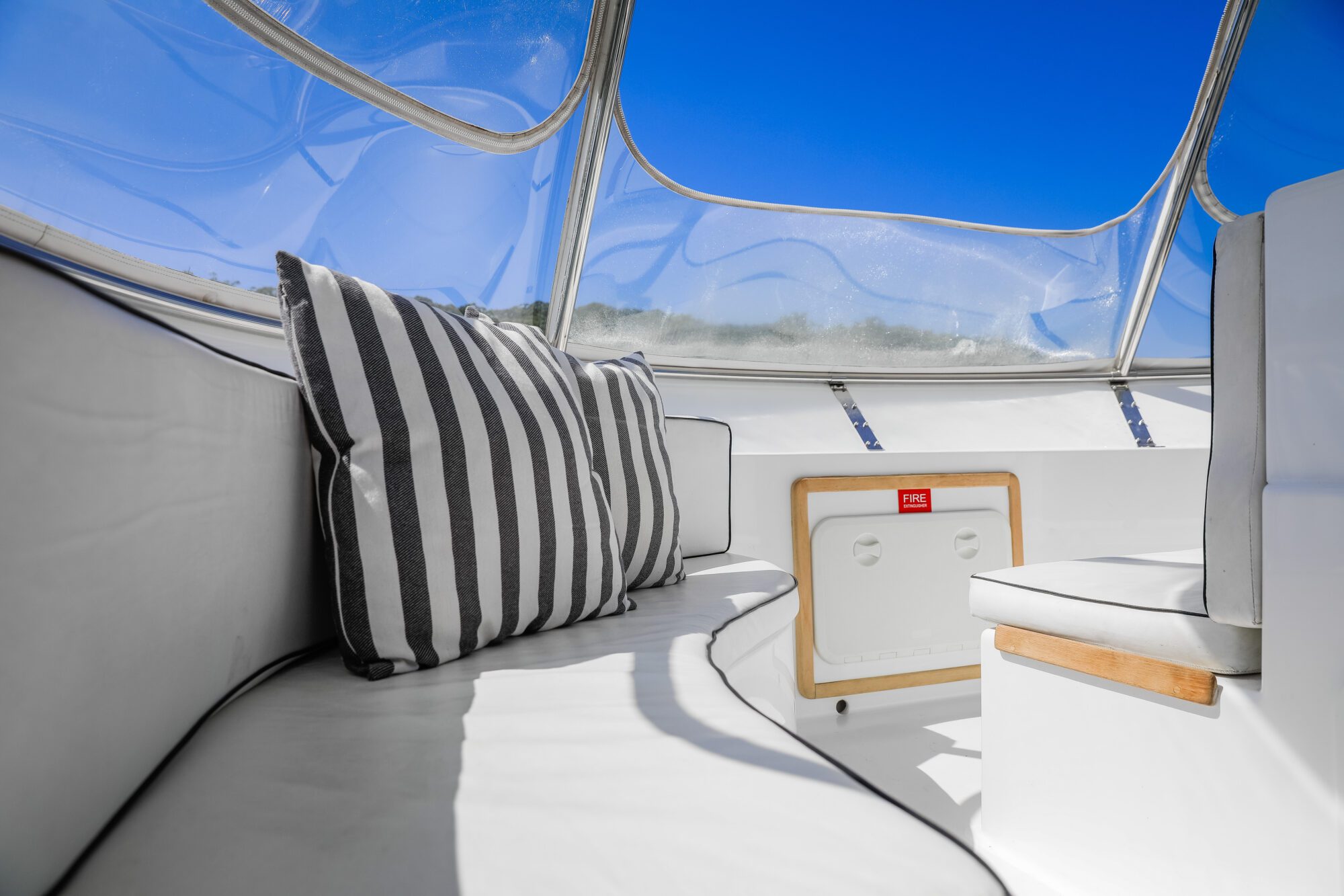 White cushioned seating area on a boat with black and white striped pillows, clear canopy cover above, and a small storage compartment. Blue sky is visible through the canopy.
