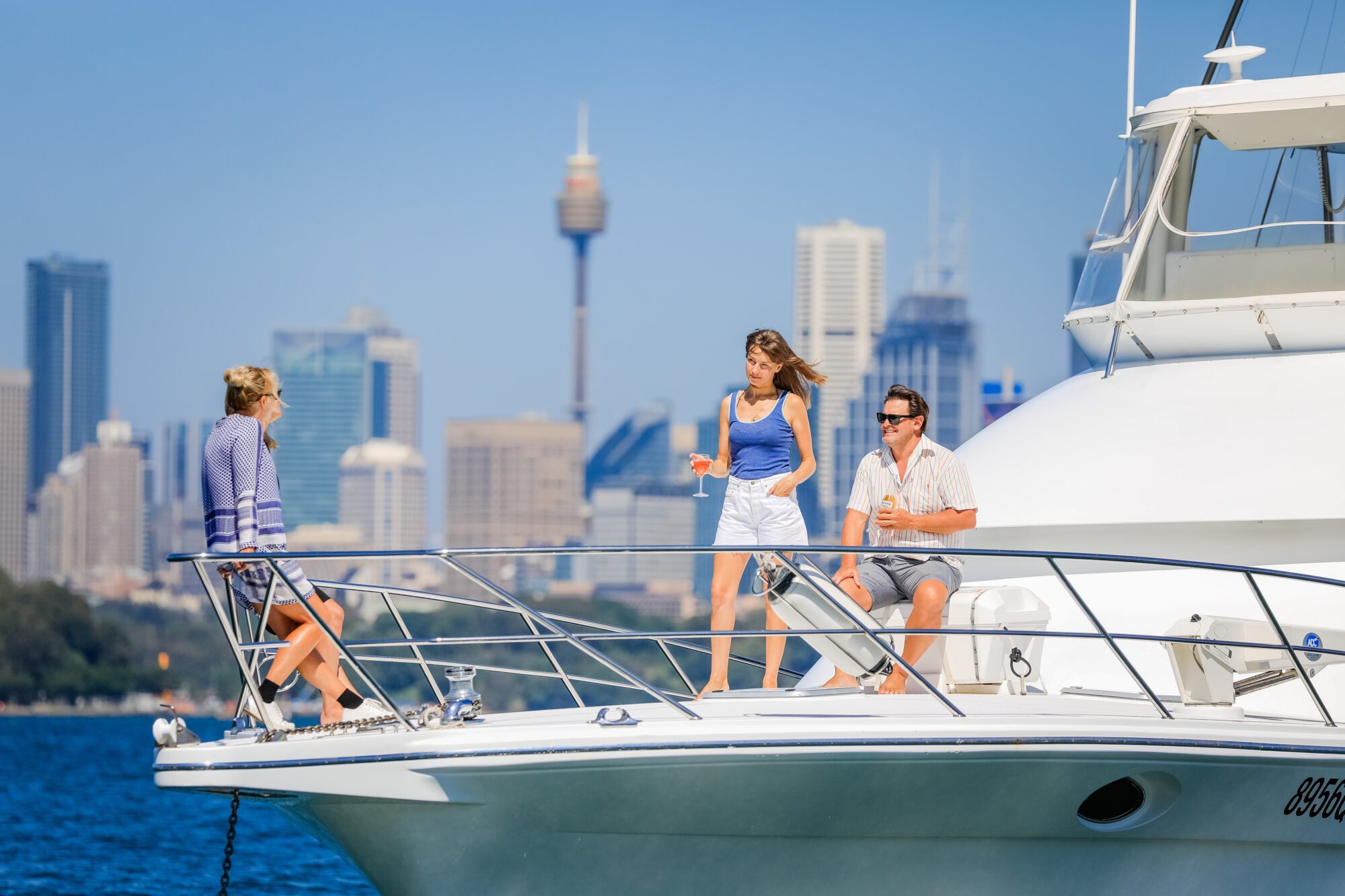 Three people relax and chat on the deck of a white yacht on a sunny day, with a city skyline and tall buildings in the background. Two are standing while one sits near the edge, all in casual summer attire.