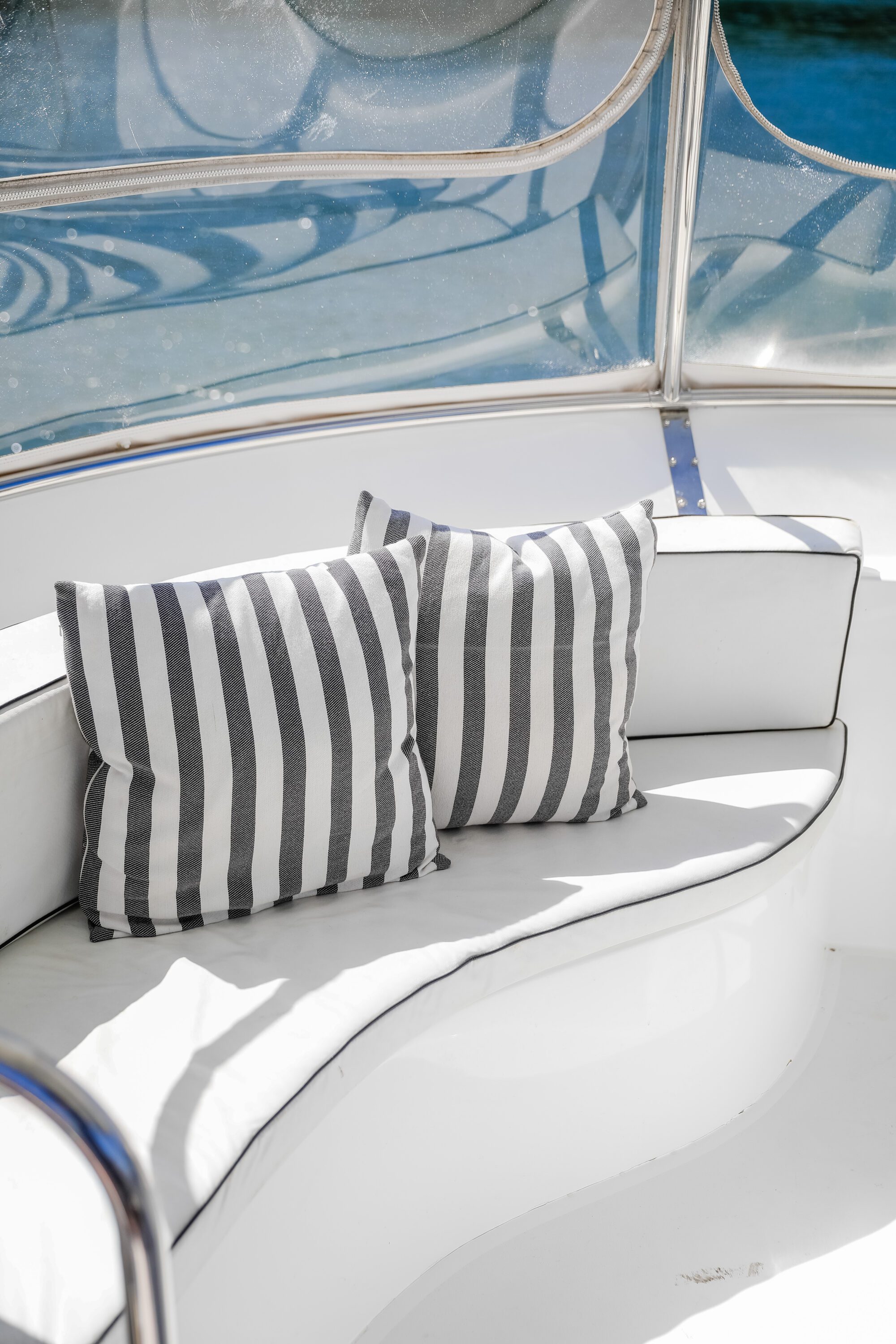 Two gray and white striped pillows rest on a curved white cushioned bench aboard a boat, with sunlight streaming in and water visible through the clear panels behind the seat.
