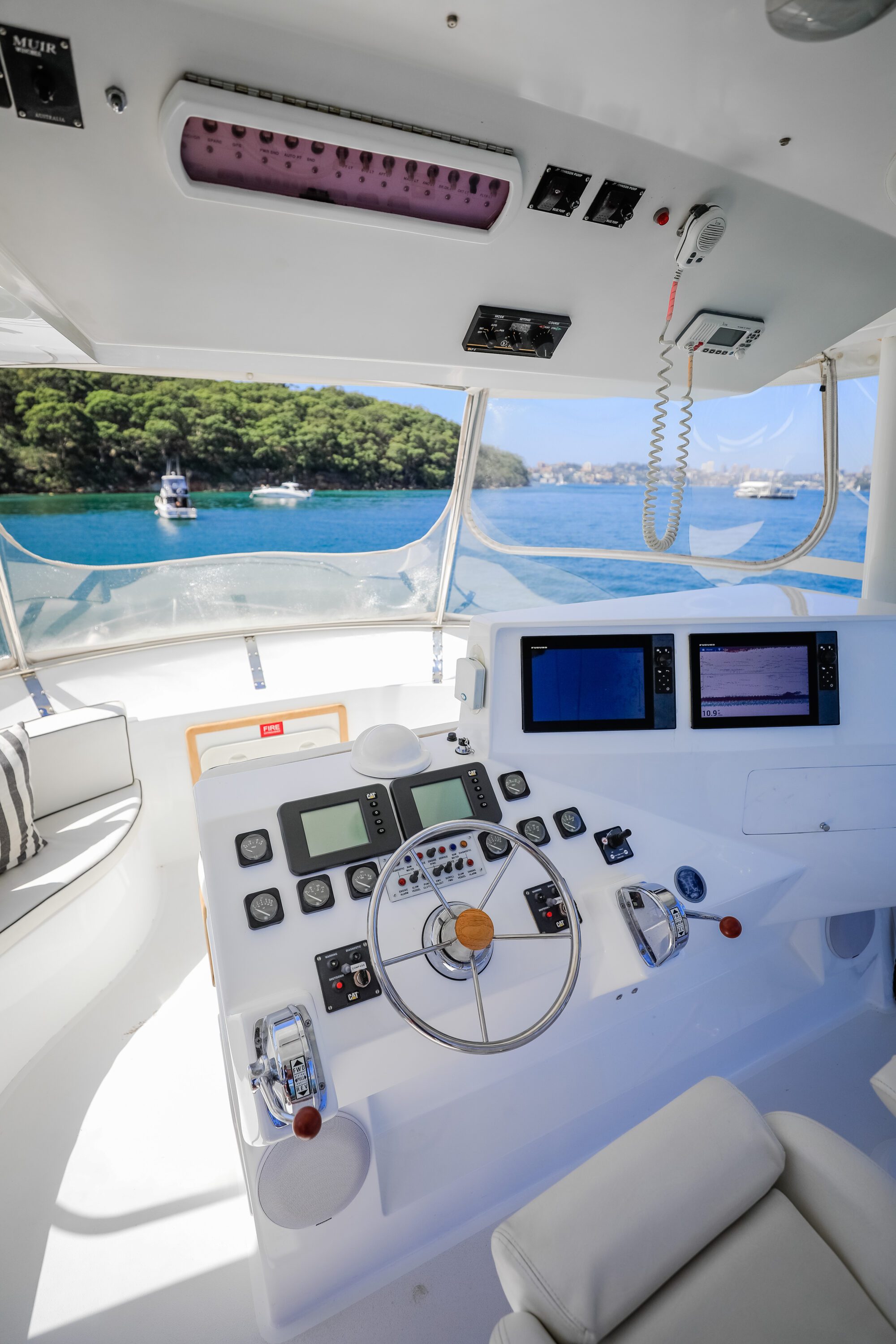 The image shows the control panel of a yacht with multiple screens, buttons, and a wooden steering wheel, overlooking calm blue water and green trees through large windows.