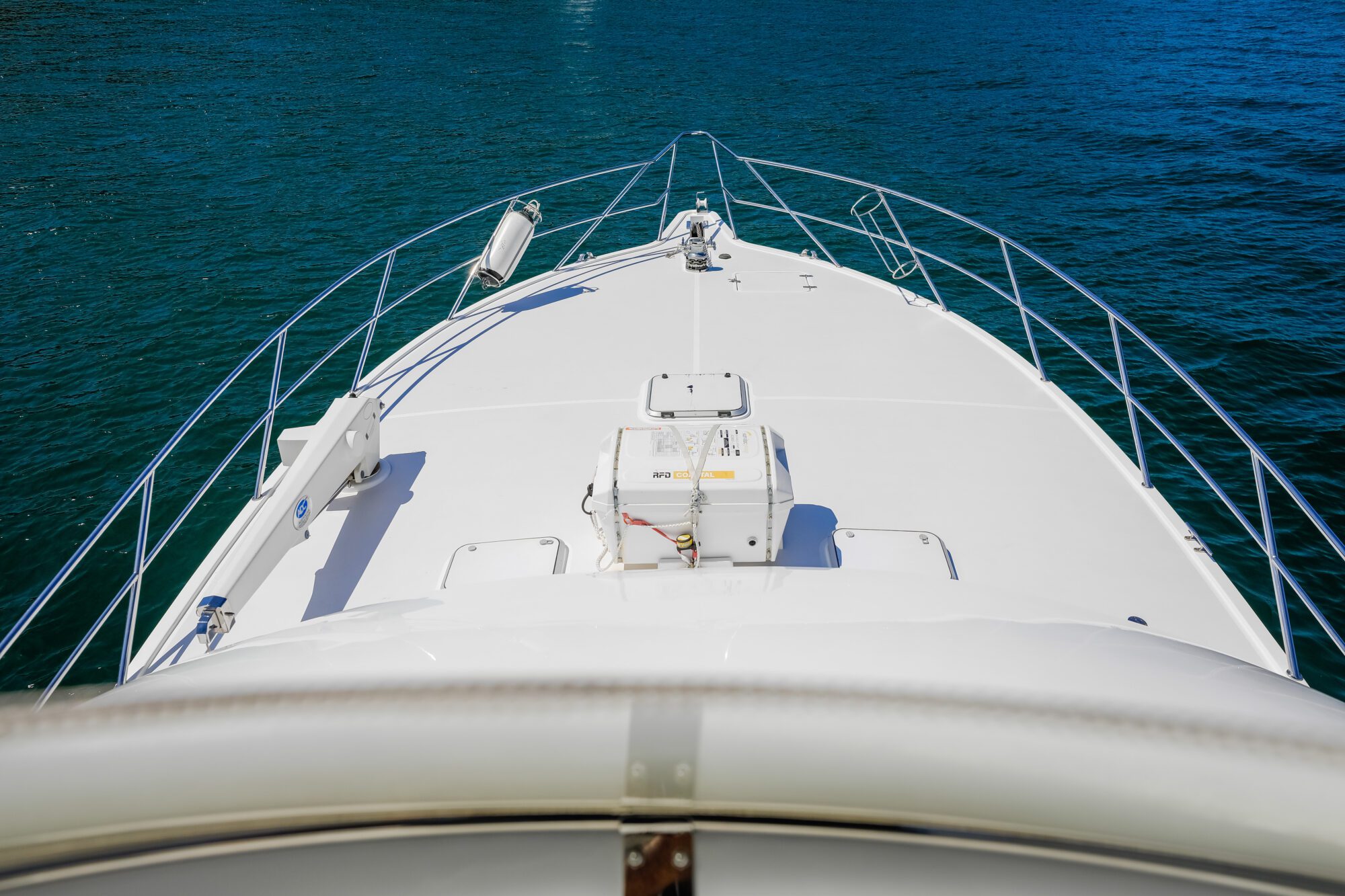 View from the bow of a white yacht sailing on calm blue-green water, showing the deck, railings, and marine equipment under a sunny, clear sky.