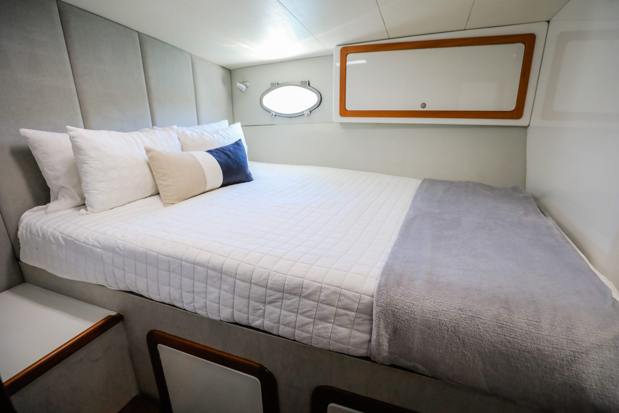 A neatly made bed with white bedding, multiple pillows, and a gray throw blanket inside a small, modern room with padded walls and a small round window.
