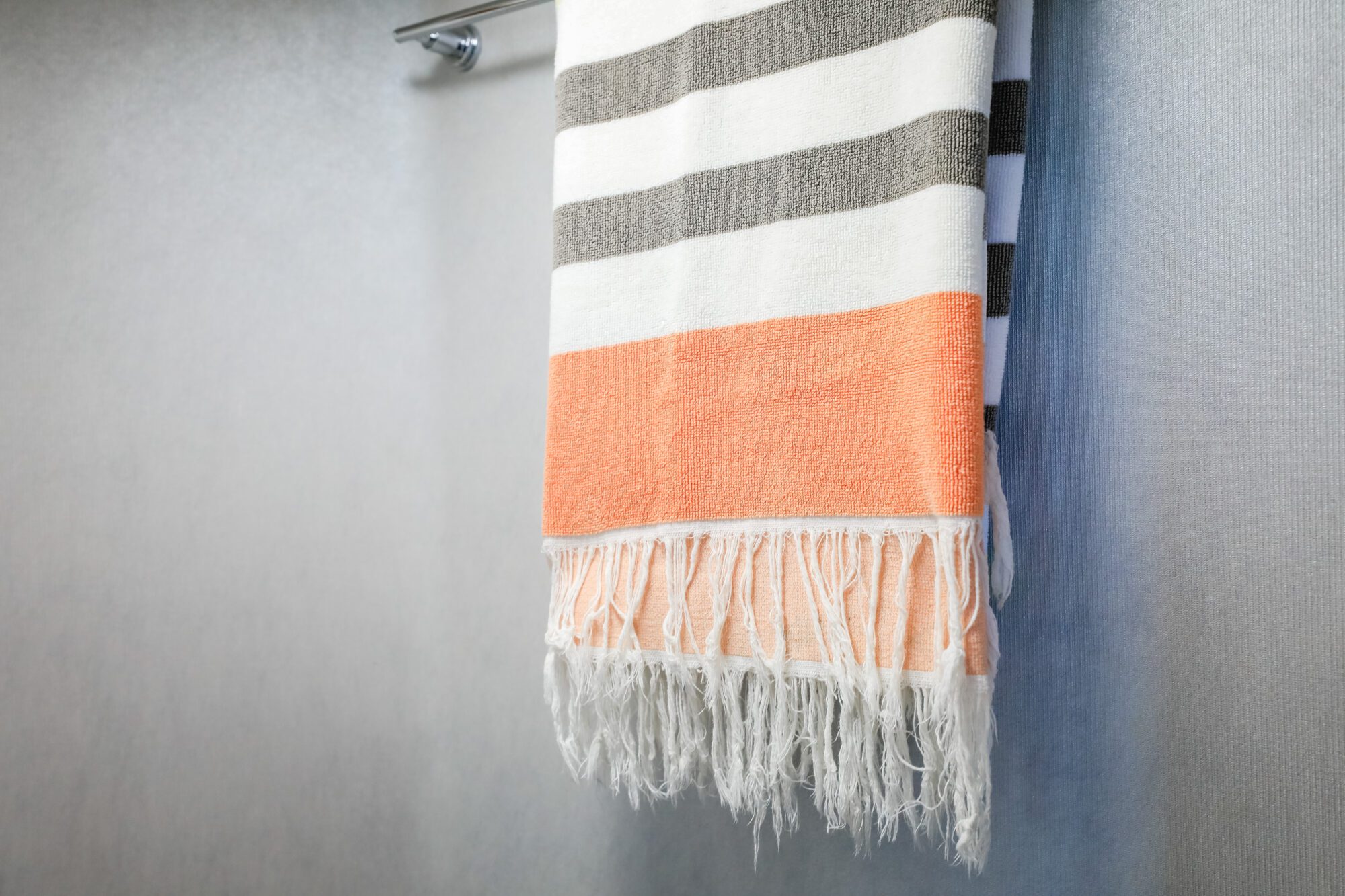 A towel with horizontal white, gray, and orange stripes and fringed ends hangs on a silver towel bar against a light gray wall.