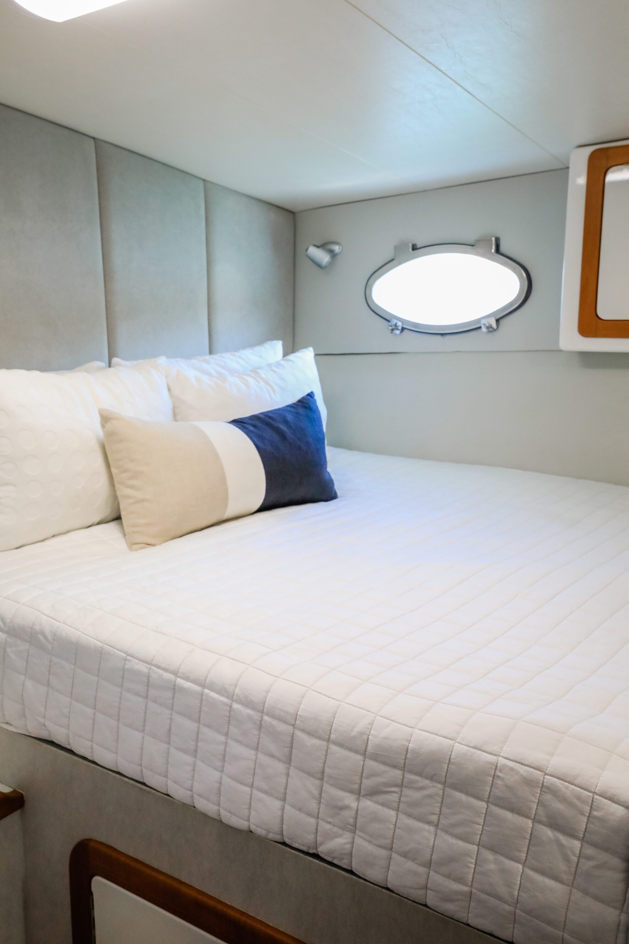 A neatly made bed with a white quilted cover and three pillows sits next to a small oval window on a boat or yacht, with beige padded walls and a wall-mounted cabinet visible.