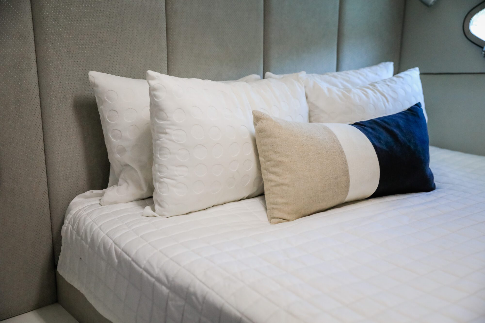 A neatly made bed with a white quilted cover, three white pillows, and a rectangular pillow with beige, white, and navy blue sections, positioned against a padded headboard.